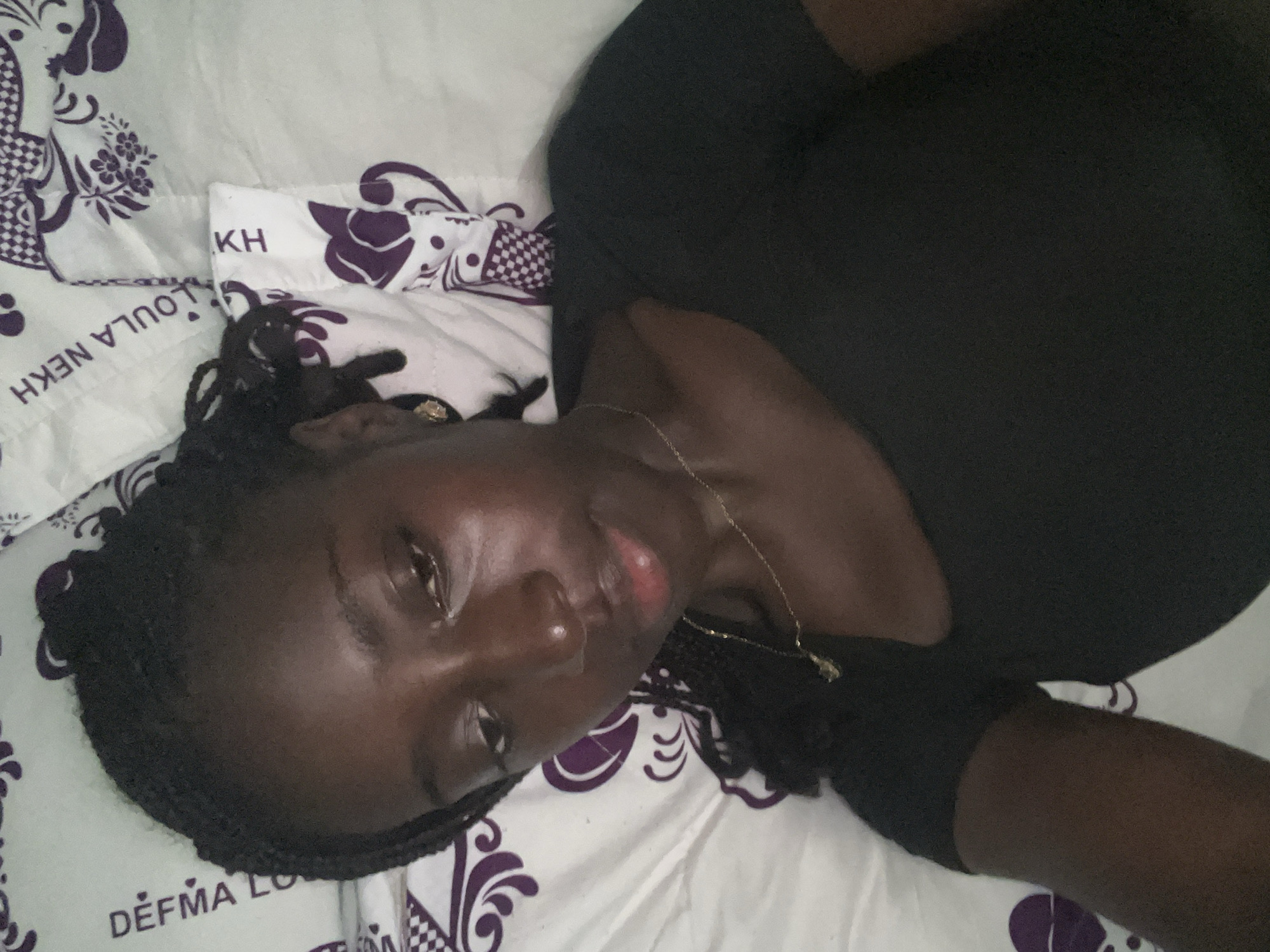 A young Black woman is lying on a bed with white and purple patterned sheets. She is wearing a black t-shirt and a gold necklace. Her hair is braided and pulled back. The sheets have various patterns, including flowers and text that appears to be in French and possibly Arabic. One section of the sheet has the text "DEFMA LO" and another has "OULA NEKH". The image is taken from a high angle, looking down at the woman. The lighting is soft, suggesting an indoor environment. There is no specific indication of the location within Abuja, Nigeria.