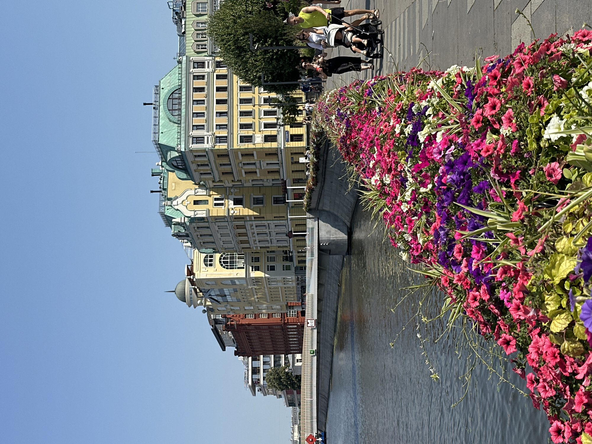 In this sunny, outdoor scene in an unknown city, a picturesque canal bank is adorned with a vibrant display of colorful petunias in shades of pink, purple, and white. The flowers form a lush border along the walkway, which runs parallel to the water. On the canal, a small red boat is visible. Across the water, a row of multi-story buildings with distinct architectural styles lines the opposite bank. The buildings vary in color, with prominent ones in red, yellow, and light green, featuring numerous windows and decorative elements. Several people are walking along the paved walkway. In the foreground, a couple with a baby in a stroller is moving away from the camera. The man is wearing a bright yellow shirt and black shorts, while the woman is in a dark dress. Further down the walkway, other individuals are seen strolling or walking. The sky is a clear, bright blue.