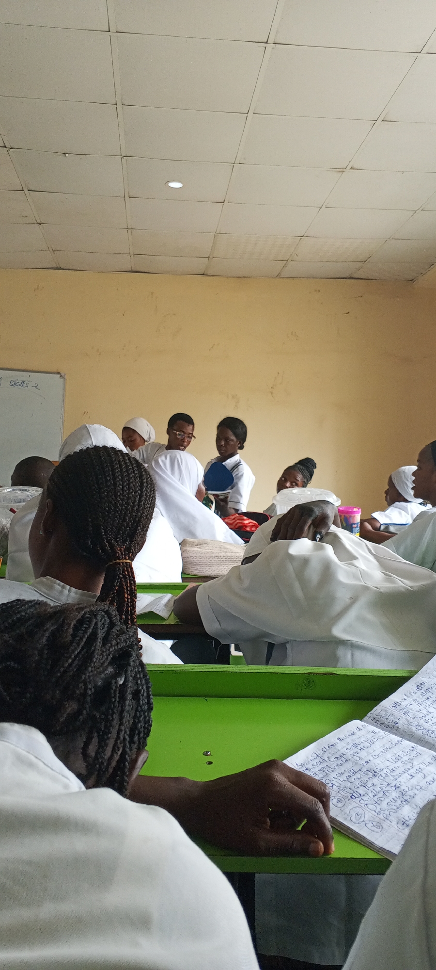 The photo shows a classroom scene in Keffi, Nigeria. A group of people, mostly women, are dressed in white uniforms, possibly nursing students. They are seated at green desks with notebooks open, suggesting a lecture or demonstration. At the front, a small group appears to be leading a lesson. The room is brightly lit, with a light colored wall and a whiteboard.