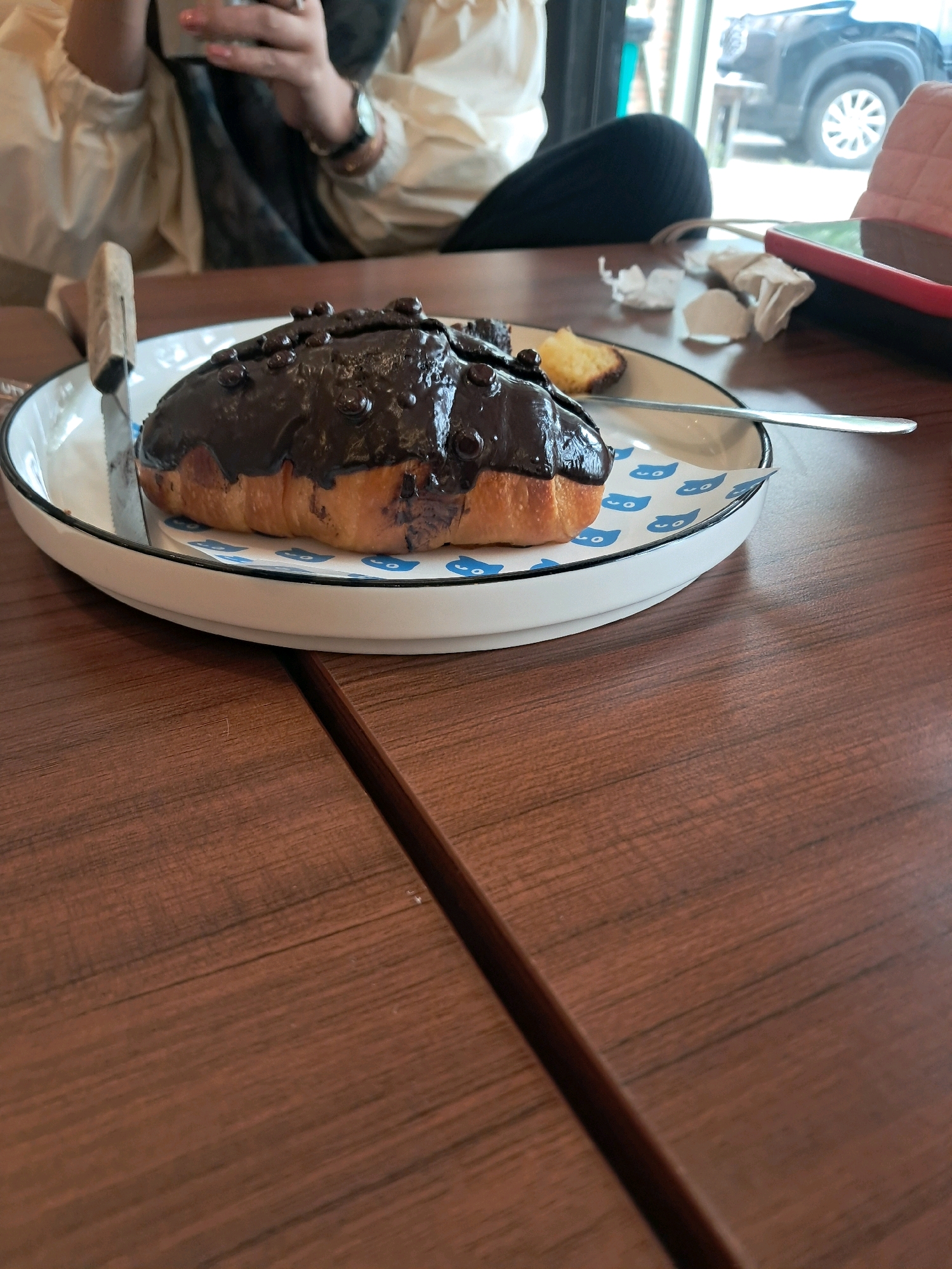 A close-up, low-angle shot focuses on a chocolate-covered pastry on a white plate with a blue pattern, placed on a wooden table. A knife with a light-colored handle is on the left side of the plate. In the background, a person wearing a light-colored blouse and dark pants is partially visible, holding a phone. Beyond the person, through a window, a black SUV is parked outside, and a pink object rests on a surface. The scene appears to be indoors, possibly at a cafe or restaurant, located in Singkawang, Indonesia.