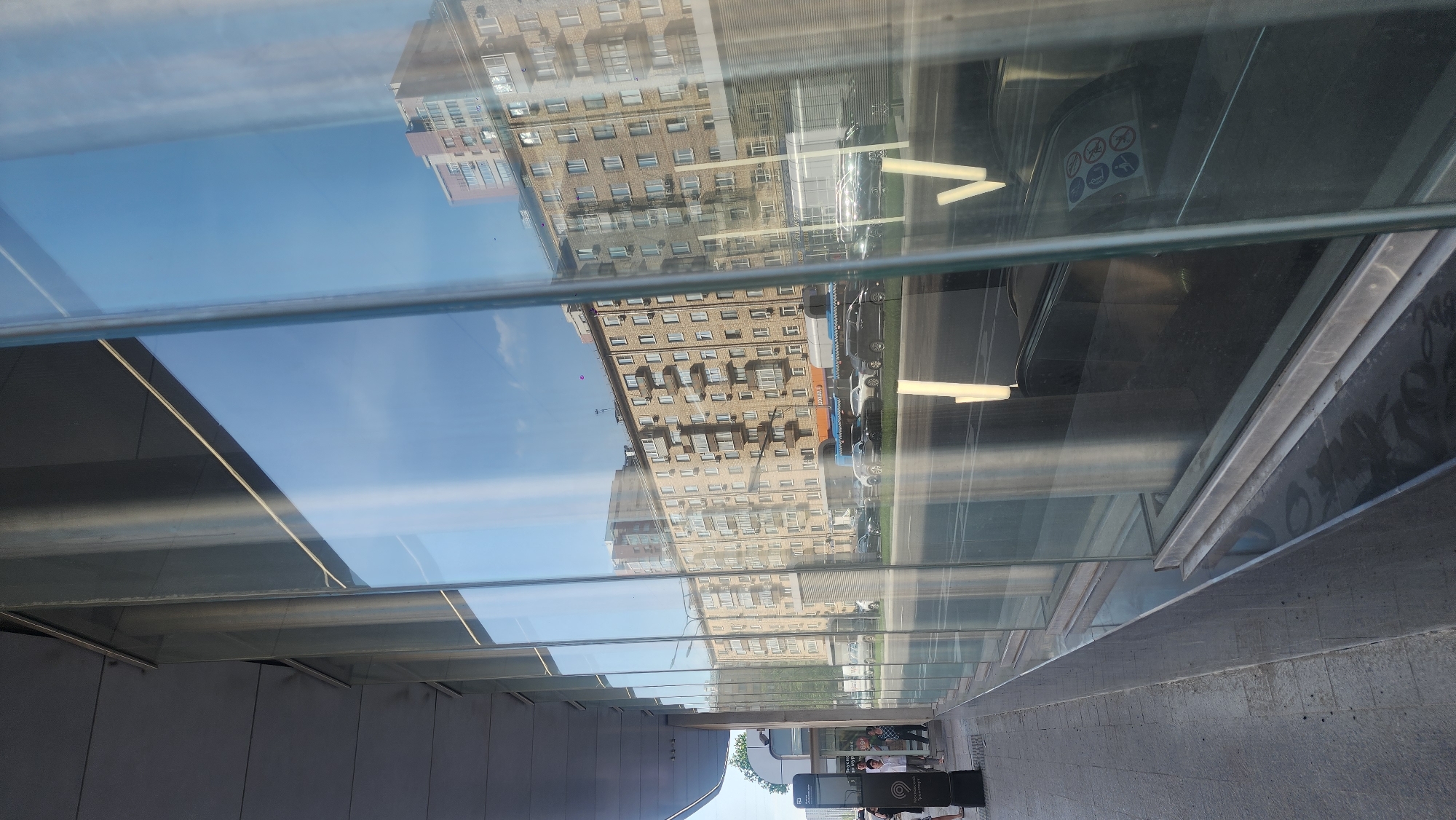 The image depicts a pedestrian underpass in Moscow, Russia, as seen through a glass ceiling. The reflection on the glass shows a multi-story apartment building across the street. Traffic, including cars and possibly buses, are visible in the reflection as well. Below the glass ceiling, people are walking through the underpass. There is a sign with the letter 'M' on it, which indicates the entrance to the Moscow Metro system.