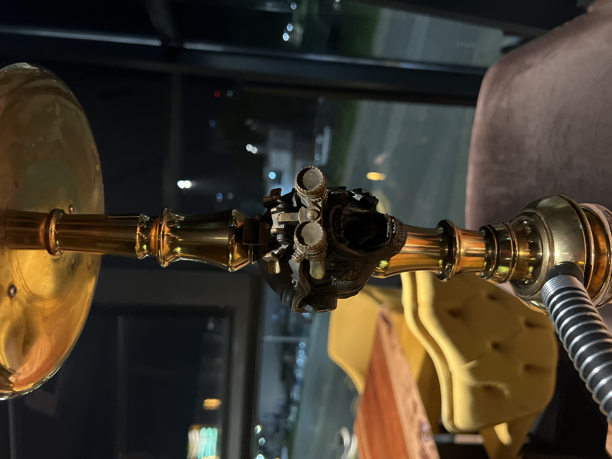 The image focuses on a brass and copper lighting fixture, possibly part of a lamp or chandelier. The fixture has intricate detailing with what appears to be a decorative piece in the center. A corrugated metal pipe is attached to the right side of the fixture. The background includes a blurred urban landscape at night, suggesting the lights of a city. Part of a chair is visible. Given the urban environment and available lighting, it is plausible that the photo was taken indoors at night in a public establishment or private residence in Burgas, Bulgaria.
