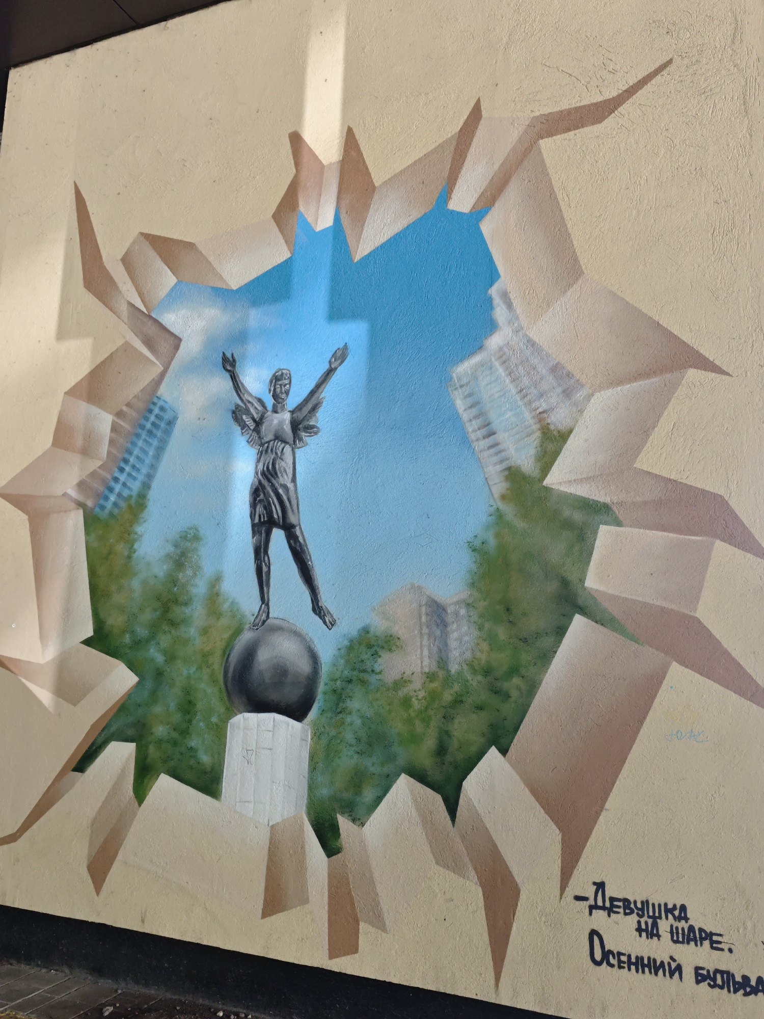 The image is a mural painted on a textured, cream-colored wall. The mural depicts a trompe-l'œil effect, simulating a hole in the wall that opens to a sky-blue expanse. Within this opening, a stylized statue of a winged figure, presumably female, stands with arms outstretched, atop a dark, spherical object, which in turn rests on a white pedestal. Behind the statue, there are painted green trees and hints of modern buildings with blue-tinted windows. The "hole" itself is rendered with sharp, angular, brown and cream-colored planes, giving the impression of shattered material. In the lower right corner of the mural, there is graffiti in black, Cyrillic lettering that reads "-Девyшka НА ШAPE. ОСEHHий БУЛЬВAР." This translates to "-Girl ON A BALL. Autumn Boulevard." This mural is located on the facade of the Moscow State University (MSU) dormitories on the Sparrow Hills (Vorobyovy Gory) in Moscow, Russia. The Sparrow Hills are a prominent landmark and viewpoint in Moscow.
