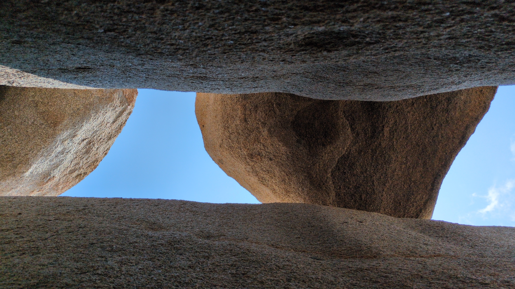 The image shows a view looking upwards through a narrow gap formed by massive rock formations. The sky is visible through the opening, appearing as a bright blue expanse with a few wisps of clouds in the lower right corner. The rocks have a rough, textured surface and are predominantly shades of brown and gray. The scene is located in Phuoc Dinh, Vietnam.