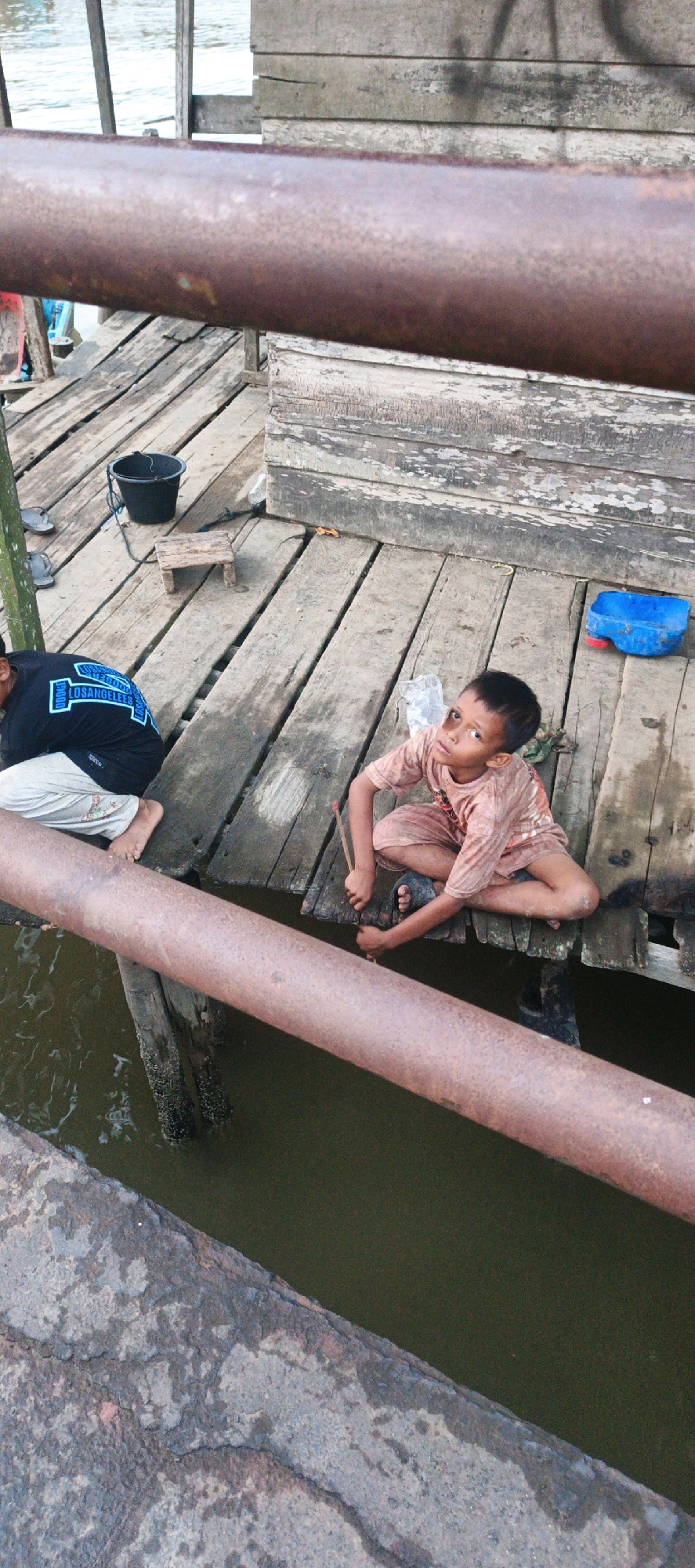 The image depicts two young boys on a weathered wooden pier, likely in Simbur Naik, Indonesia. The pier is constructed of uneven wooden planks, with some gaps revealing murky water below. A rusty, thick metal pipe runs horizontally across the foreground, partially obscuring the view.
The boy on the right is seated cross-legged, wearing a light-colored, possibly dirt-stained t-shirt and shorts. He is looking towards the camera with a curious expression. In his right hand, he holds a slender stick or twig. He is wearing dark-colored sandals.
To the left, another boy is visible from the waist down, also sitting on the pier. He is wearing a black t-shirt with a blue graphic that appears to say "LOS ANGELES" and light-colored pants. His bare feet are visible.
Scattered on the pier are various objects: a black bucket with a rope handle, a small wooden stool, a blue plastic container with a red lid, and some crumpled white plastic. The background consists of a rustic wooden wall. The environment appears damp and worn, suggesting an outdoor, possibly rural or coastal, setting.