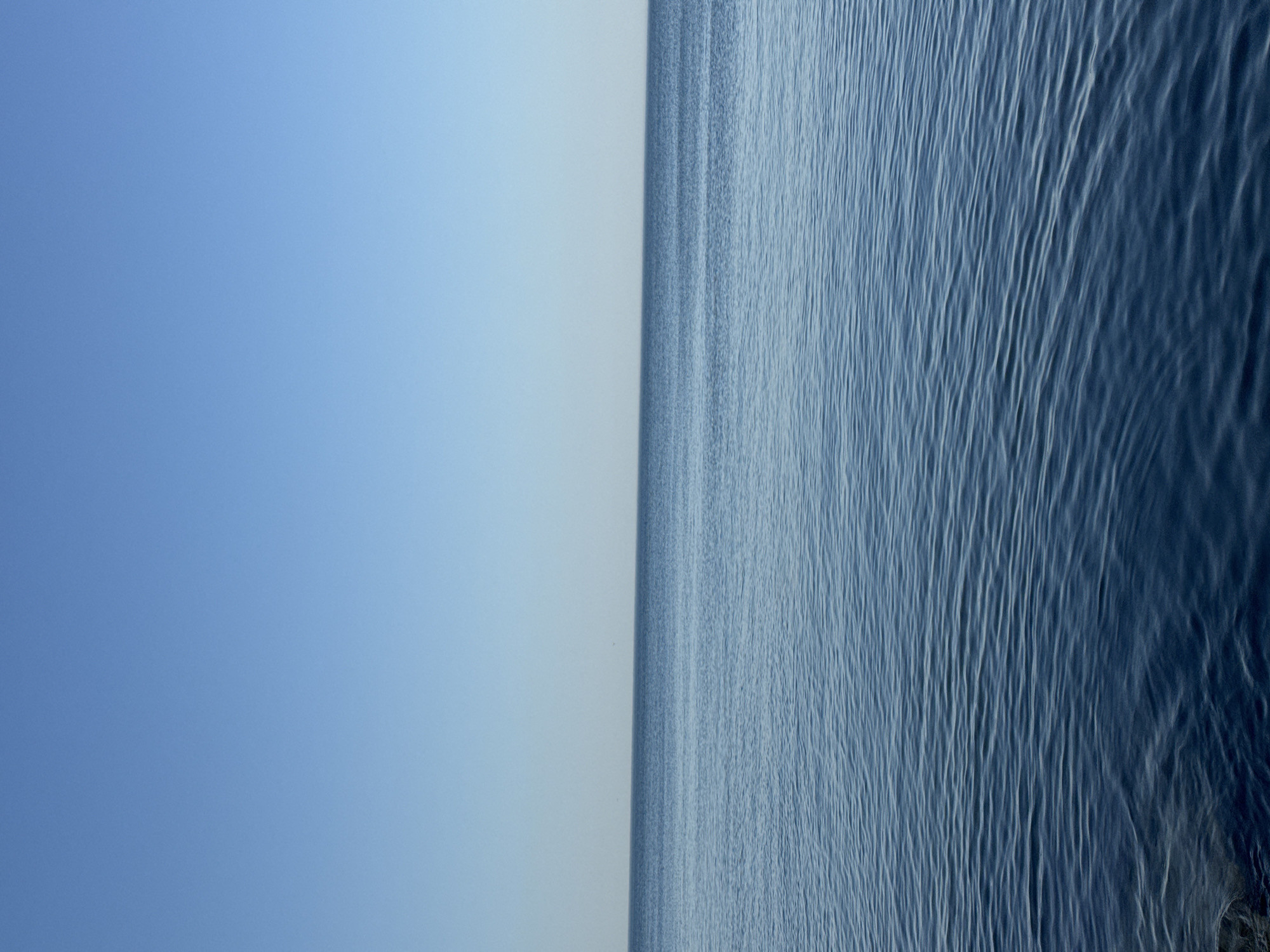 The image depicts a vast expanse of calm, blue water meeting a clear, bright blue sky at the horizon. The water exhibits gentle ripples, suggesting a light breeze. The scene is entirely natural, with no man-made structures or human presence visible. The location is identified as the Caspian Sea shoreline in Aktau, Kazakhstan. The overall impression is one of serene natural beauty and tranquility, characteristic of a coastal landscape.
