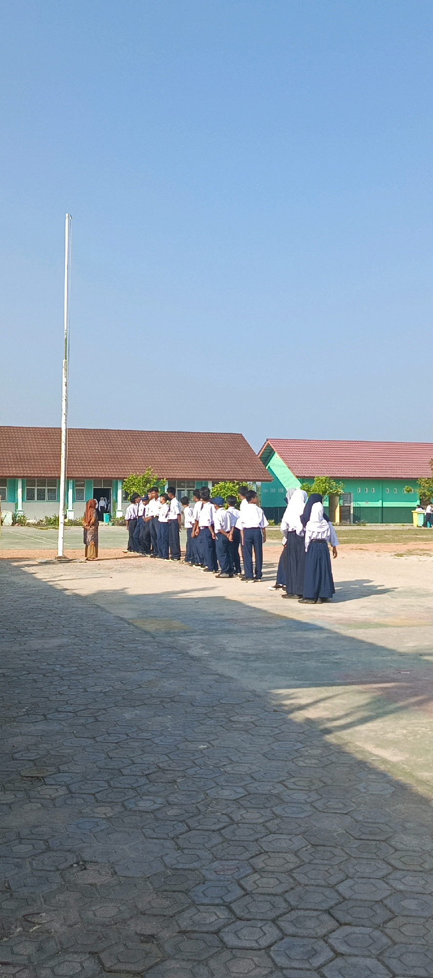 In Desa Sarang Mandi, Indonesia, a group of students in uniform are lined up outdoors on a paved area in front of school buildings. The students, some in white shirts and dark pants, and others in white shirts and dark skirts with headscarves, are facing away from the camera. One adult, possibly a teacher, is standing to the left of the student line, facing them. A flagpole stands to the left of the frame. The school buildings have reddish-brown tiled roofs and light-colored walls. The sky is clear and blue.