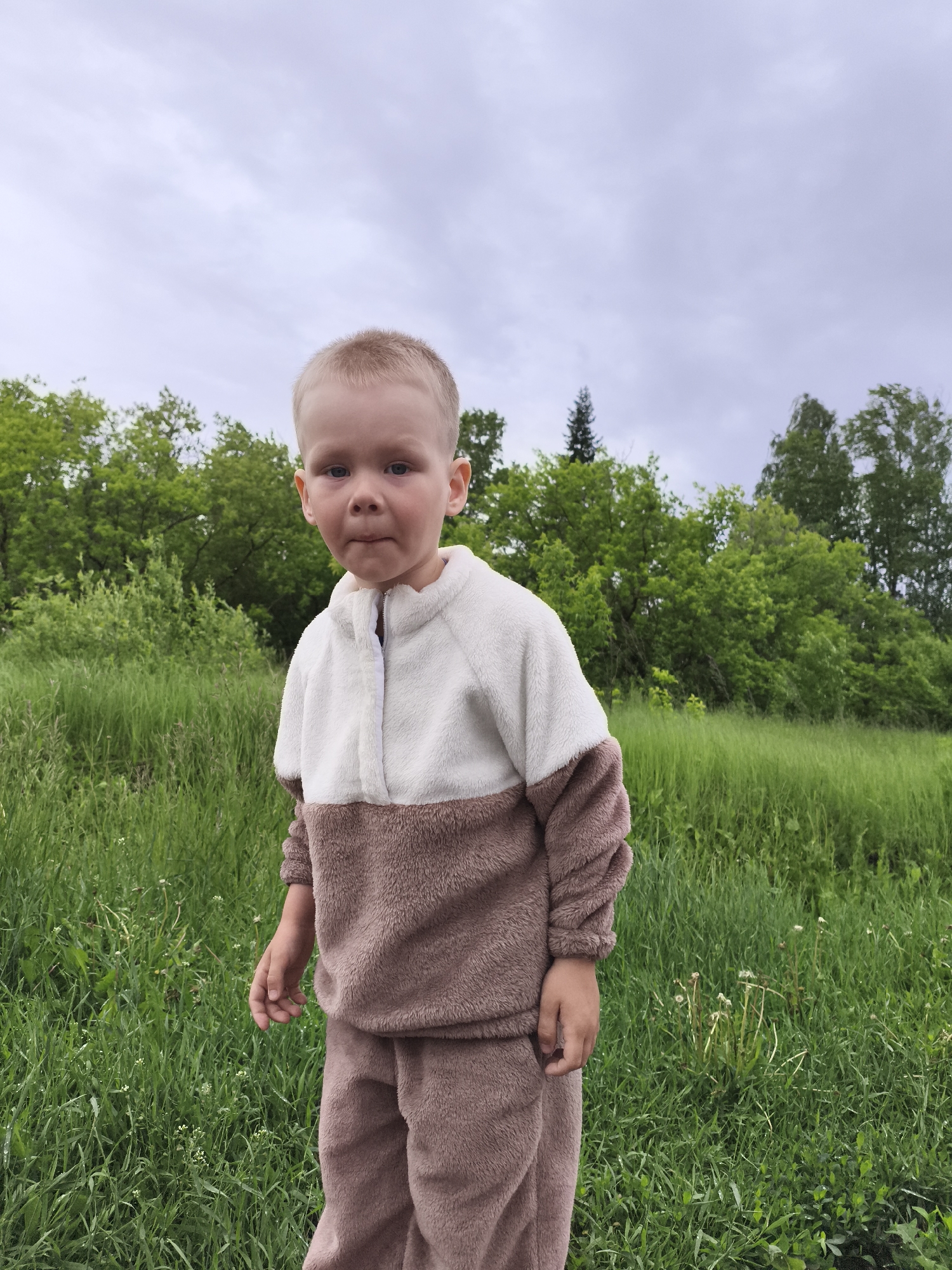 The image depicts a young child, approximately 2-4 years old, standing outdoors in a grassy field in the city of Можга, Russia. The child, with fair skin and short, light-colored hair, is positioned centrally in the frame, looking directly towards the camera with a neutral expression, exhibiting slightly pursed lips. The child is wearing a two-piece fuzzy fleece tracksuit: the upper portion of the top is off-white, while the lower portion of the top, sleeves, and pants are a uniform light brown or taupe. The top features a short zippered placket at the neckline.
The immediate foreground consists of dense, verdant green grass with scattered small white wildflowers and some dried plant stalks. Behind the child, a field of tall green grass and lush green foliage of various bushes and trees extends into the midground and background. A discernible line of deciduous trees forms the main treeline, with one taller, darker evergreen tree visible to the left within the tree line. The sky above is overcast and light grey, indicating cloudy weather. The overall setting is natural and rural, dominated by green vegetation under a muted sky.