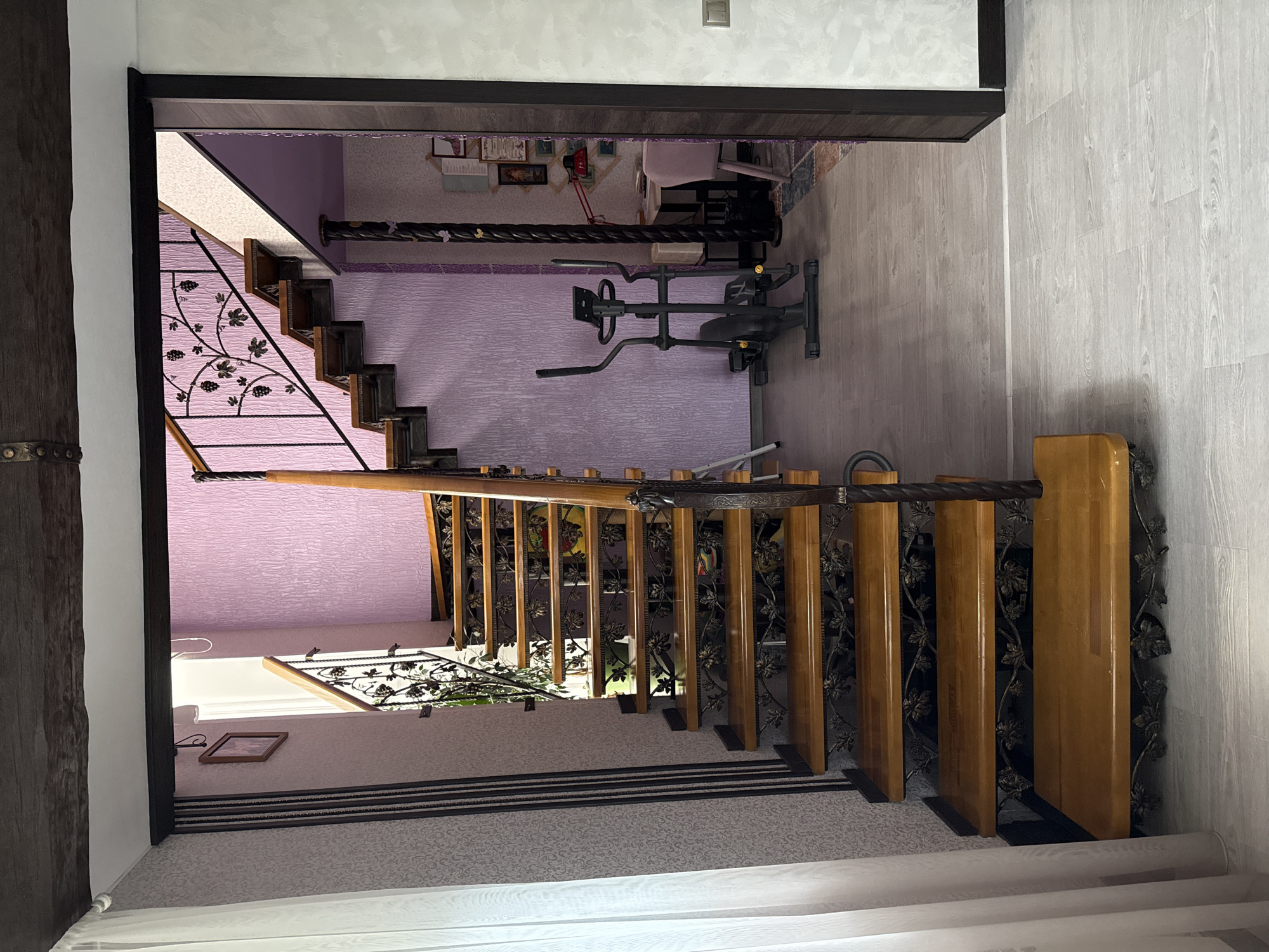 This media file presents an interior view of a multi-level home in Krapivka, Russia, focusing on a prominent staircase descending from an upper floor. The staircase features light-colored wooden treads and risers, complemented by a striking black wrought-iron railing adorned with intricate grape and leaf motifs, topped with a dark wooden handrail.
At the upper landing, a stationary exercise bike, primarily black and gray, is positioned on light wood-look flooring. The walls in this upper area are covered with a textured, crinkled purple wallpaper. Beyond the exercise bike, in an adjacent room, framed pictures are visible on the wall, along with a small red desk lamp and hints of furniture, possibly a chair or desk.
At the bottom of the visible stairs, a landing is covered with a light-colored carpet featuring a subtle patterned design. A dark wooden beam is visible on the far left, and another section of the ornate railing extends further down out of frame. Light wood-look flooring also appears on the right side of the lower area.
The scene is well-lit, suggesting daytime. No people are present, and there is no visible text. The overall aesthetic combines traditional elements, like the ornate staircase, with modern household items like the exercise equipment.