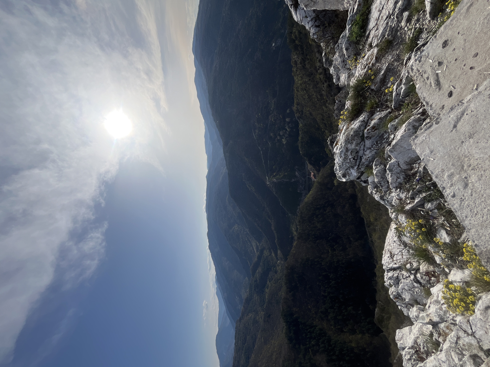 The image is a landscape photograph taken from a high vantage point, possibly a cliff or mountain edge. The foreground features rocky terrain with some vegetation, including small yellow flowers. A steep drop-off suggests a significant elevation change.
In the background, a series of mountain ridges extend into the distance, creating a layered effect. The sky is visible, with a bright sun and some clouds. The lighting suggests it is daytime.
The location might be in or near Asenovgrad, Bulgaria, given the mountainous terrain characteristic of the region. However, specific landmarks or features would be needed to confirm the exact location.