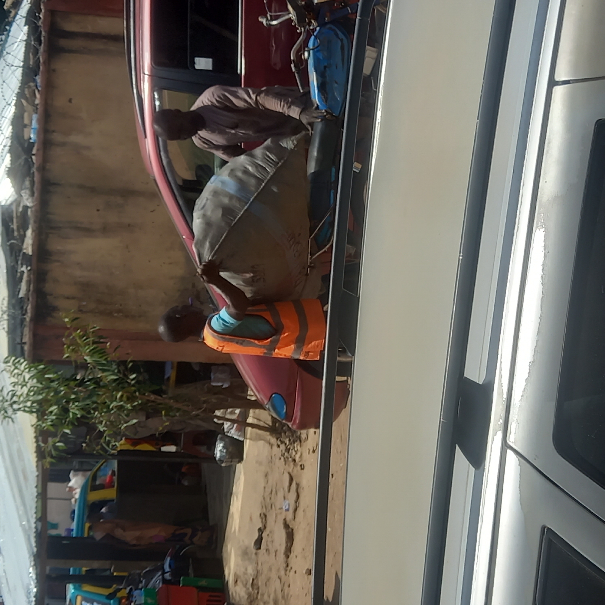 In Sardauna, Nigeria, two men load a red vehicle. One man, wearing a light-colored shirt, is inside the car near a blue container. The other, wearing a blue shirt and orange safety vest, carries a large sack towards the vehicle. Various objects and vehicles are in the background. A building stands nearby, with a shelter-like structure visible in the lower left corner. The ground appears sandy and uneven. A white vehicle is prominently featured in the foreground.