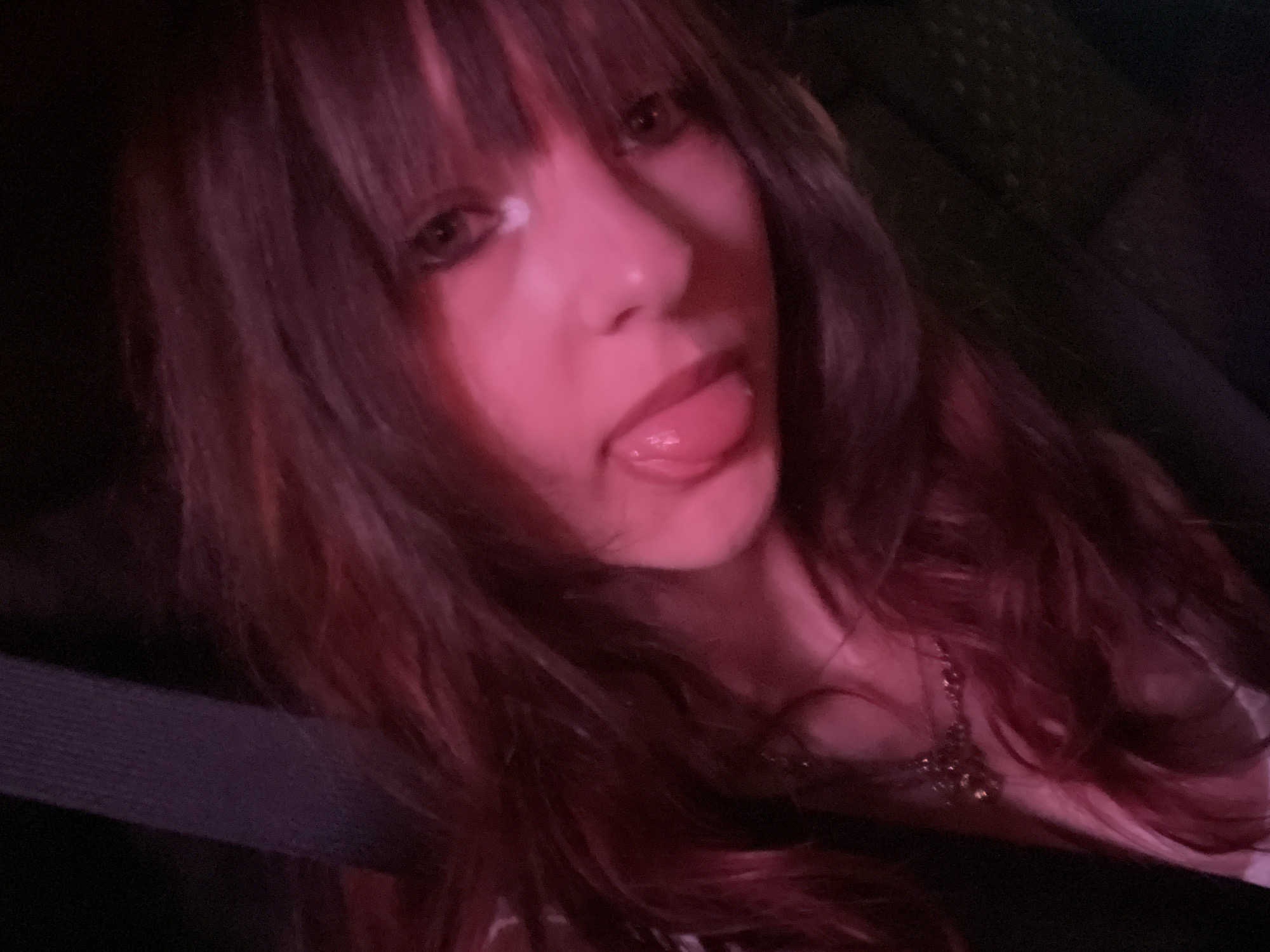 The image is a close-up, angled selfie of a young person with dark, layered hair, partially illuminated by pinkish light. The person is sticking their tongue out, with their eyes open and looking towards the camera. A silver necklace with a dark pendant is visible around their neck. The background is dark and out of focus, with a hint of a textured surface, possibly a seatbelt, on the left. There is no discernible location in the city of Bnei Brak, Israel.