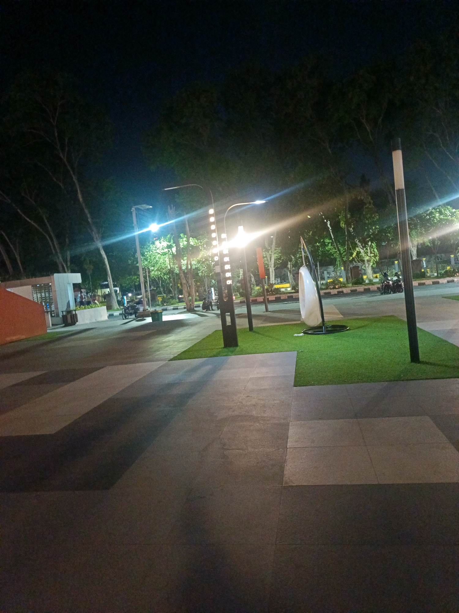 The image depicts a nighttime scene in Sidoarjo, Indonesia, likely in a public park or recreational area. The foreground is dominated by a paved walkway with large rectangular tiles in alternating shades of light gray and dark gray, creating a geometric pattern. Shadows from overhead lights and trees are cast across the walkway. To the right, a section of artificial turf grass is visible, bordered by the walkway.
In the middle ground, several elements are present. A modern, white, egg-shaped swing seat is suspended from a dark metal frame on the artificial turf. Behind this, there is a paved area with a street running parallel to it, where several motorcycles are parked. A person is pushing a handcart on this paved area. Further back, a building with a prominent white "T" shaped structure is visible. This building appears to be a part of a larger complex, with some people gathered near its entrance.
The background features a dense arrangement of trees, their branches silhouetted against the dark night sky. Multiple streetlights are illuminated, casting bright beams and creating lens flare effects in the image. One prominent streetlight is a tall, dark pole with a white top. Another is a curved lamp post with multiple lights arranged vertically. The overall atmosphere is one of a well-lit, accessible public space at night.