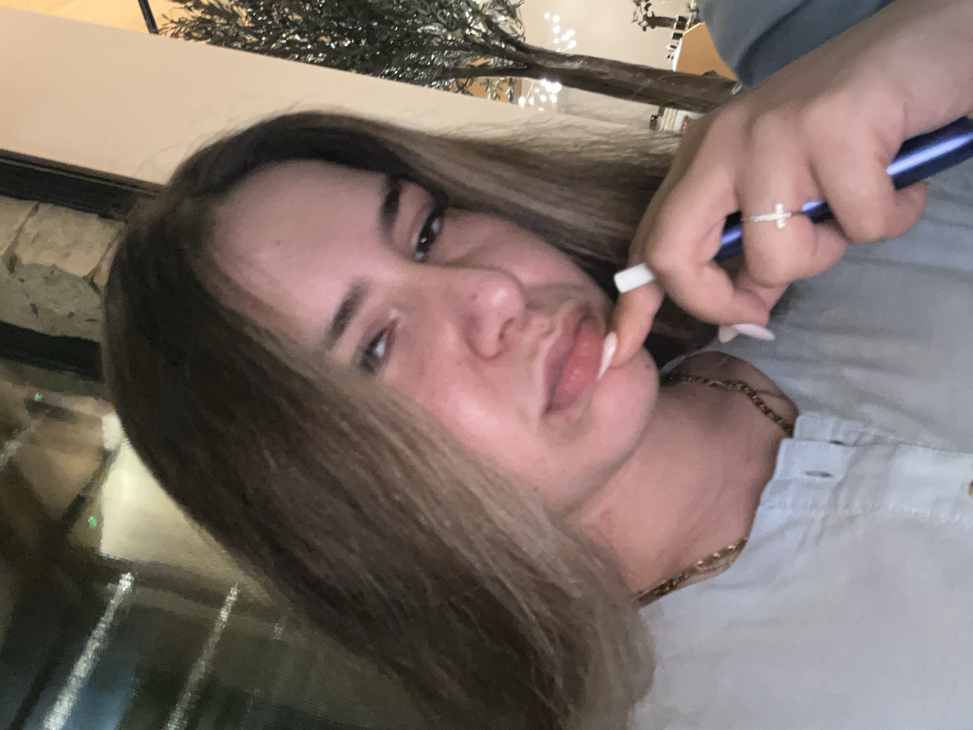 A young woman with long, light brown hair is pictured from the chest up, looking downwards and to her left. She is wearing a light blue collared shirt and a gold chain necklace. Her right hand is near her face, with her index finger touching her chin and her thumb holding a blue pen. She is wearing a silver ring with a cross on her right ring finger and has long, light-colored fingernails. The background is dimly lit, with a blurred reflection of lights and what appears to be an olive tree on the right side. The setting suggests an indoor location, possibly a restaurant or cafe, in Nicosia, Cyprus.