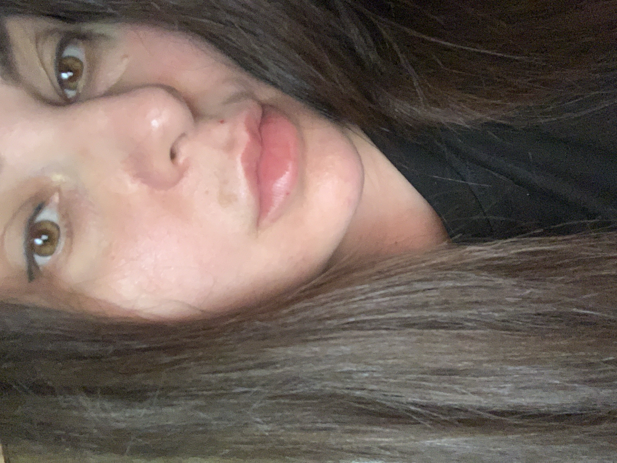 The media file is a close-up, sideways portrait of a person's face. The subject has brown eyes, a nose, and lips. Their skin appears to have a rosy hue, with some visible texture. Long, dark brown hair surrounds the face, framing it and obscuring parts of the background. The hair is styled in loose waves. The lighting is soft, creating subtle highlights on the skin and hair. The overall impression is one of a relaxed, natural pose, possibly taken indoors due to the lighting. There is no visible text or any specific location cues. The emotional tone is neutral.