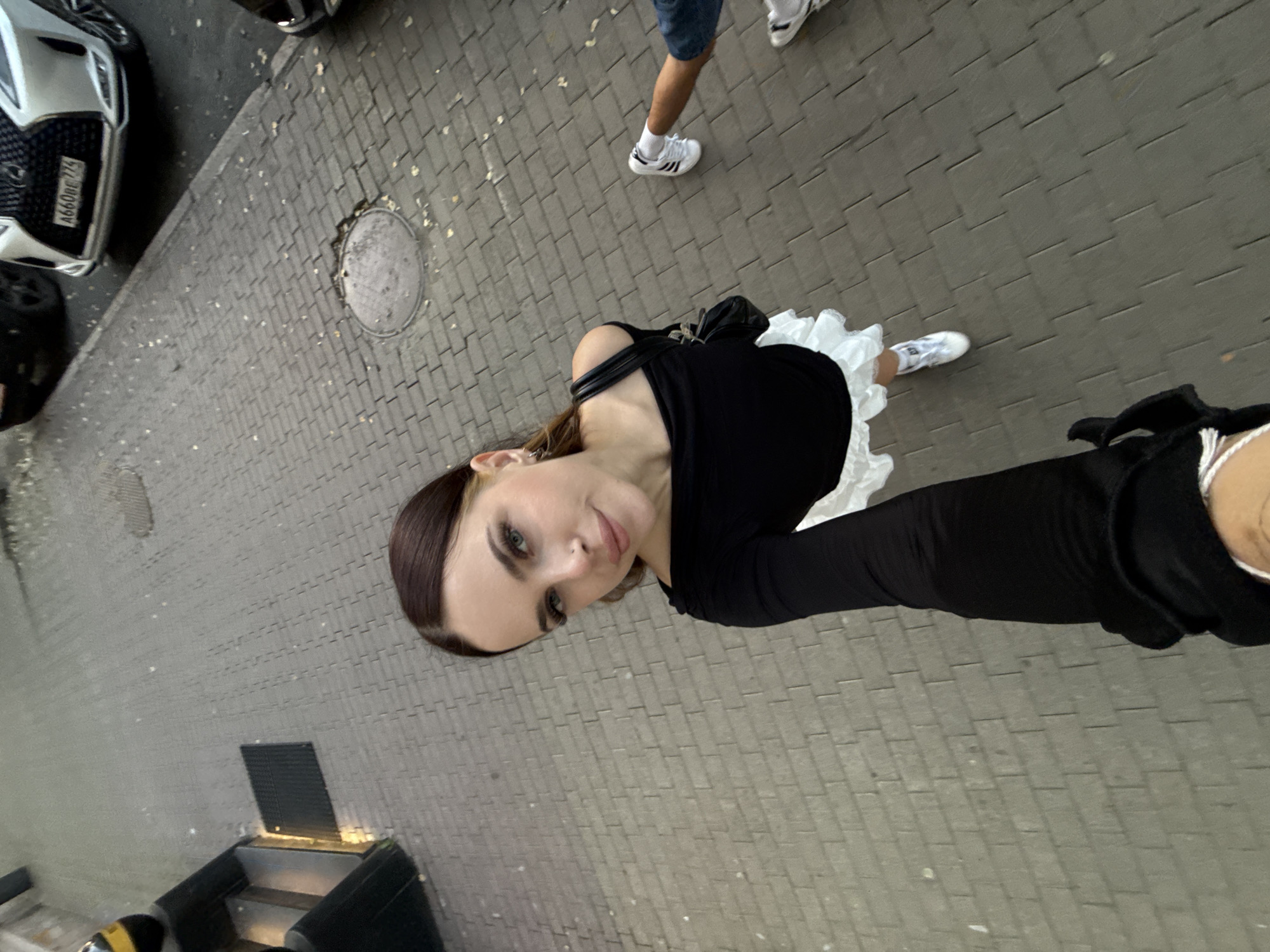 A young Caucasian woman with brown hair and blue eyes is taking a selfie outdoors on a paved sidewalk. She is wearing a black off-the-shoulder top with a white ruffled skirt and white sneakers. A black handbag is slung over her shoulder. The sidewalk is made of grey brick pavers, and there are fallen leaves scattered on the ground. In the background, a white Lexus car with a Russian license plate is visible, parked on the street. The scene appears to be in a city environment, likely Chelyabinsk, Russia, given the license plate style.