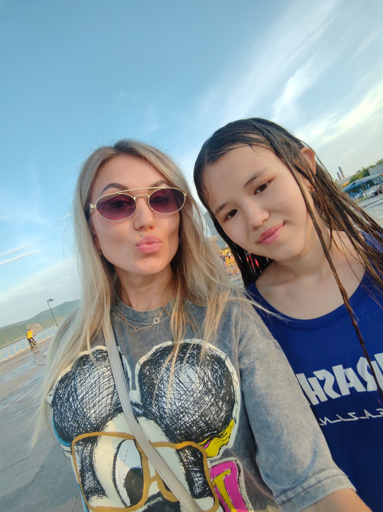 A selfie is taken by two females outdoors with a cloudy sky overhead. The female on the left has blonde hair and is wearing sunglasses and a gray t-shirt with a graphic print. She is puckering her lips. The female on the right has dark, wet hair and is wearing a blue t-shirt. They appear to be standing on a walkway with a body of water and a distant city skyline in the background. The location is not specified.