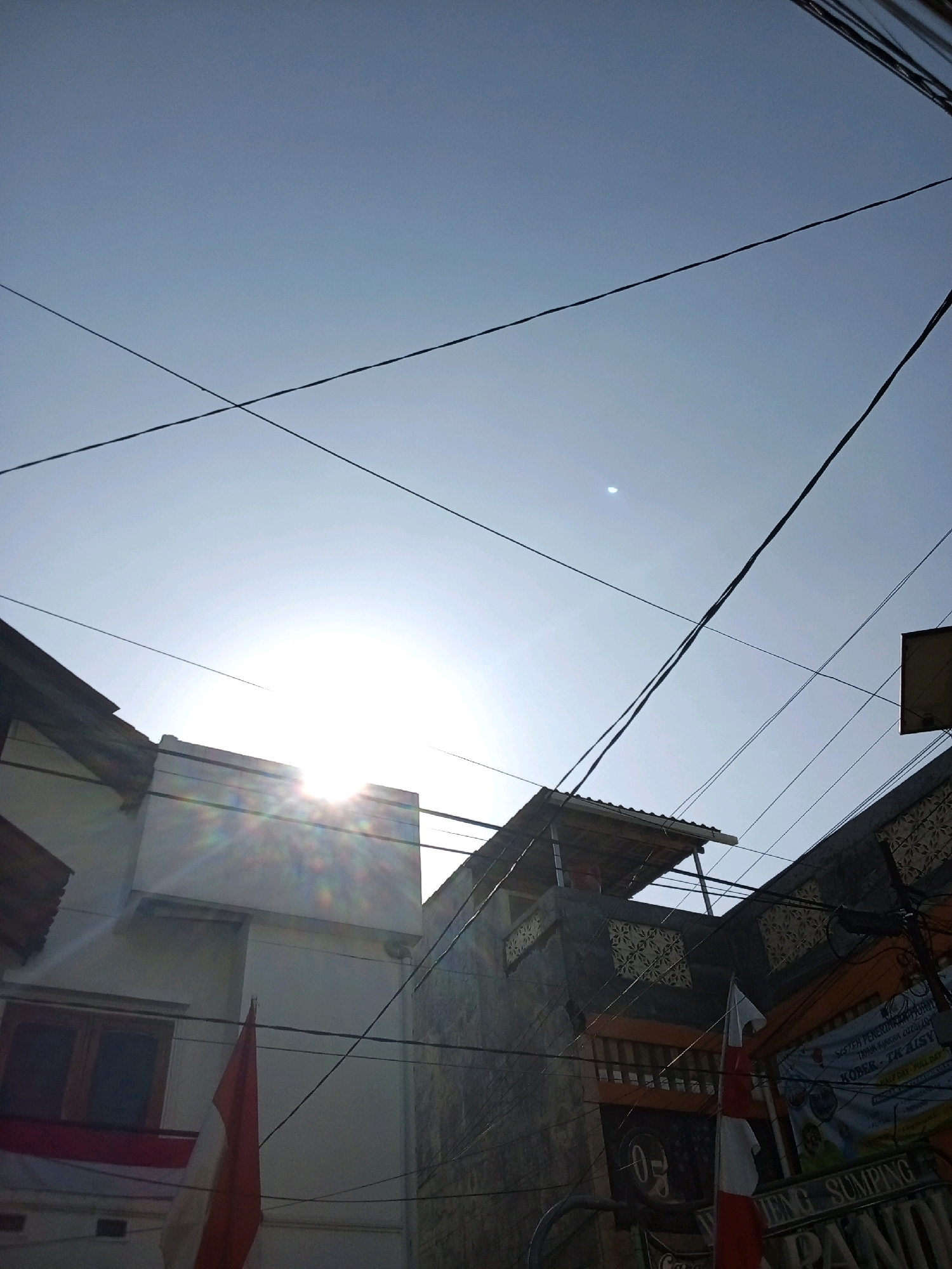 The image shows a street scene in Bandung City, Indonesia, looking up towards the sky. The sky is clear and bright with the sun shining through, causing a lens flare effect. Several electrical wires crisscross the sky, creating a cluttered visual. In the foreground, there are parts of buildings, including a white wall with a window and a small section of an Indonesian flag hanging. Another building to the right has a more textured facade and a roofline. A banner is partially visible on the building to the right, displaying some text. The overall impression is of a typical urban environment with overhead power lines and residential or commercial buildings.