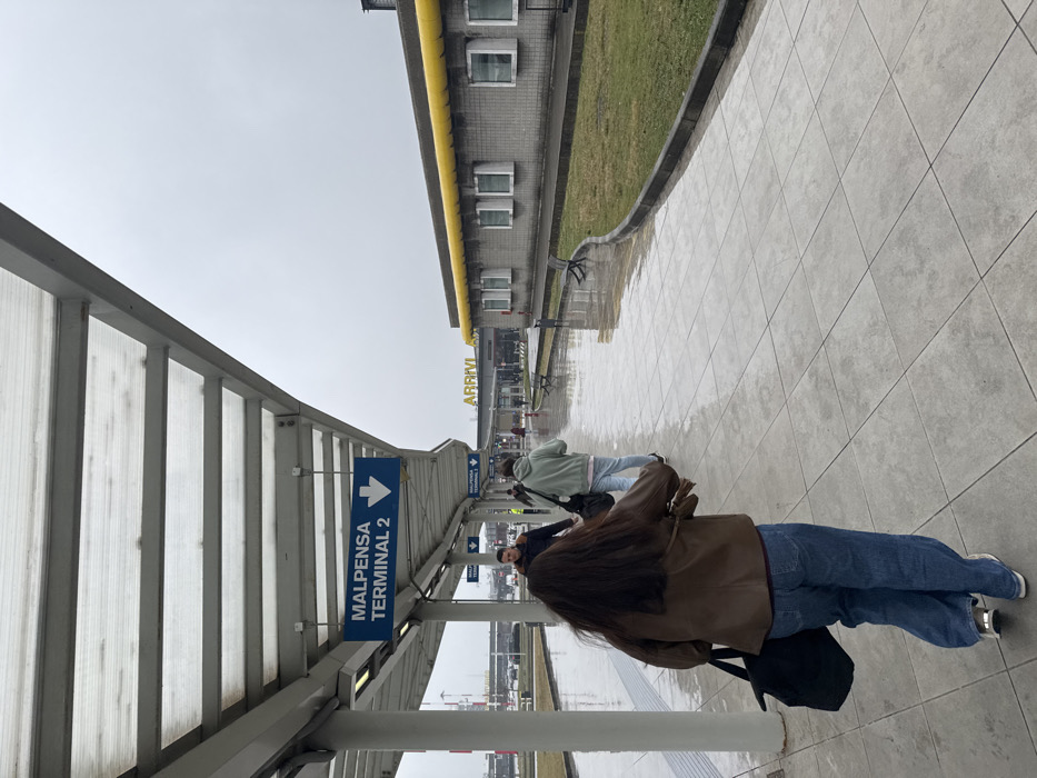 This image depicts an outdoor pedestrian pathway at Milan Malpensa Airport, Terminal 2, located near Somma Lombardo, Italy. The scene is observed under an overcast sky with a wet, light gray tiled ground, indicating recent precipitation.
A translucent corrugated canopy with a white metal framework covers the pathway. Multiple blue directional signs hang from the canopy, displaying "MALPENSA TERMINAL 2" with a downward-pointing white arrow.
In the foreground, a person with long, dark brown hair, wearing a brown leather jacket, blue jeans, and light-colored sneakers, walks away from the camera. This individual carries a black bag in their left hand. Immediately ahead, a second person in a light green hooded sweatshirt, light-colored pants, and sneakers walks in the same direction, carrying a dark bag over their right shoulder. Further down the path, a male figure wearing a dark jacket walks towards the camera. Additional indistinct figures are visible in the background, walking along the path.
To the right of the pathway, a strip of green grass separates it from a single-story, light gray building. This building features a distinctive bright yellow roofline or gutter system and multiple rectangular windows with white frames. The word "ARRIVI" in yellow capital letters is prominently displayed on a darker section of the building's facade in the background, indicating the arrivals area. Several dark benches are situated on the grassy strip. Beyond the building, airport infrastructure and vehicles are visible under the uniformly gray sky.