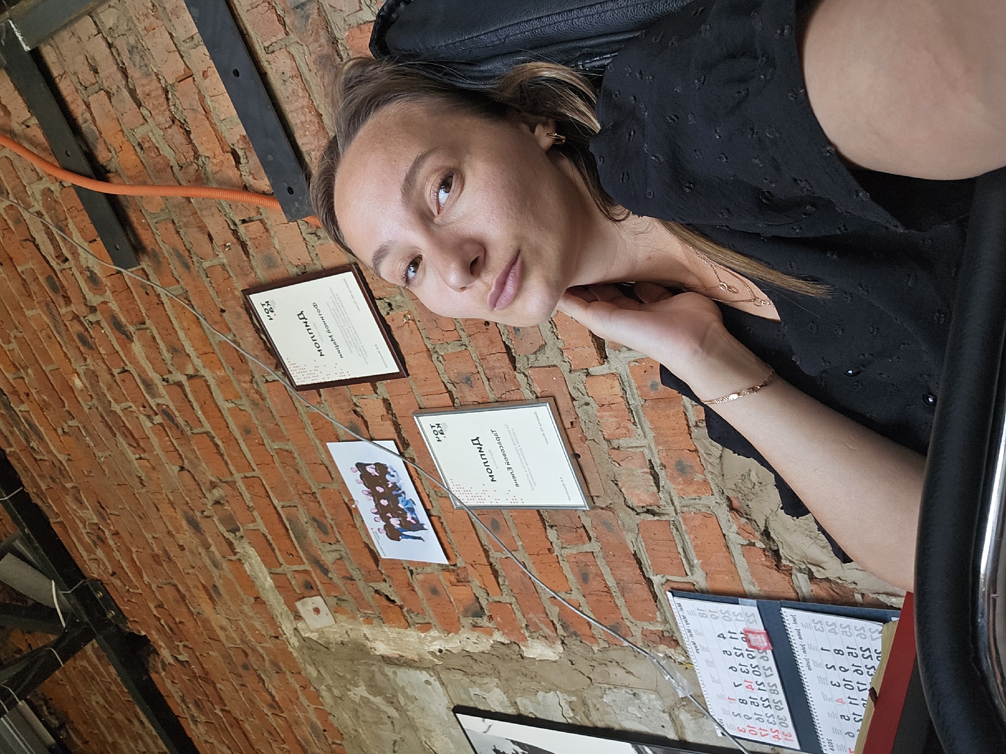 The image is a selfie of a woman with light brown hair, wearing a dark, textured top and a gold necklace. She has fair skin and is looking at the camera with a slightly pensive expression. Her right hand is near her neck.
The background consists of a weathered brick wall with various items attached to it. These include: two framed certificates or documents with text in Russian, a picture of figures with possibly balloons, and a calendar. There's also a black metal structure and an orange cable running along the wall. The presence of the calendar and documents suggests an office or work environment.
Given the OCR text includes "Чайковский" (Chaikovsky), it can be concluded that the location of this image is Chaikovsky, Russia.