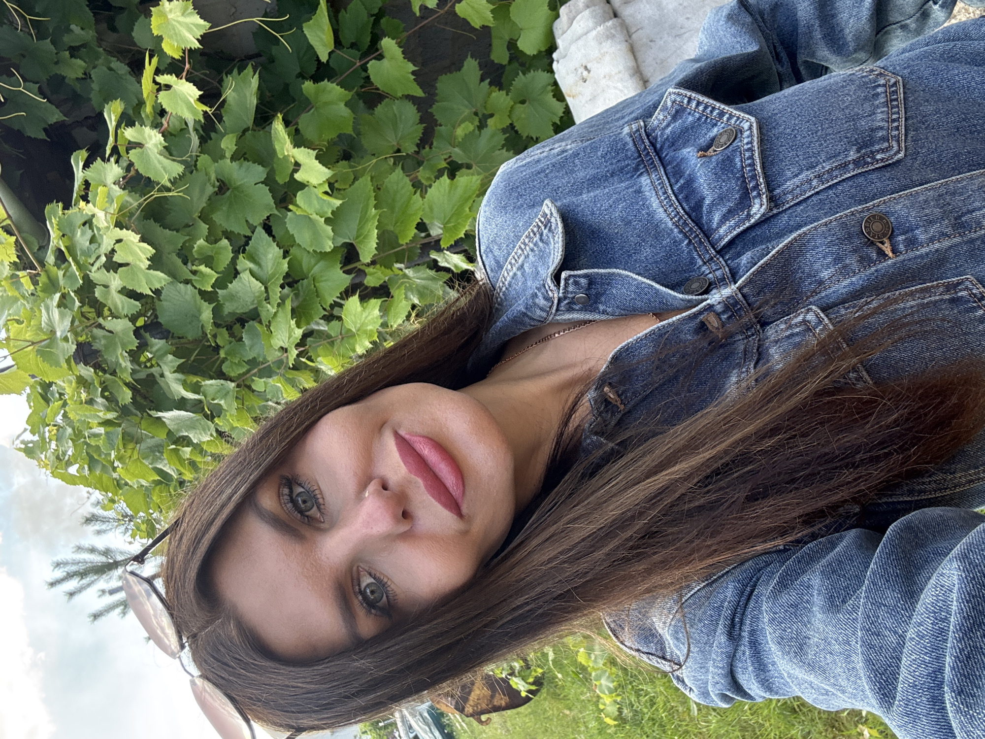 A woman with long, dark brown hair and light blue eyes is taking a selfie outdoors. She is wearing a denim jacket and sunglasses are perched on top of her head. She has on pink lipstick and is wearing a delicate necklace. In the background, there are green leaves and grass. The location is specified as Dubovaya, Russia. The overall scene suggests a casual, sunny day.