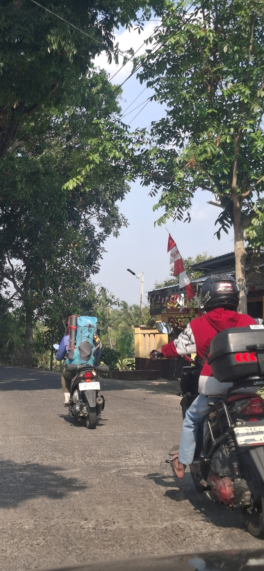 Two individuals are riding motorcycles on a paved road. The motorcycle in the foreground is black with a large black storage box on the back. The rider is wearing a red jacket, blue jeans, and a black helmet with a white logo. The motorcycle in the background is also black and is being ridden by a person wearing a blue shirt and carrying a large blue backpack with red straps. The road is flanked by trees and foliage, with a building and an Indonesian flag visible in the distance. The sky is clear and blue. The image appears to have been taken on a sunny day. The location is identified as Cibinong Hilir, Indonesia.