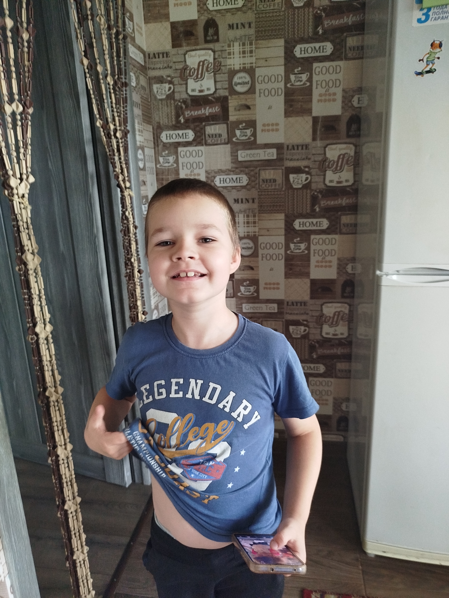 A young boy with short brown hair and a bright smile is standing in a room, holding up the hem of his blue t-shirt with his left hand. The t-shirt has "LEGENDARY" and "COLLEGE" written on it, along with stars and graphics. He is also holding a smartphone in his right hand, which is resting on his lap.
The boy is positioned in front of a wall decorated with a repeating pattern of coffee-themed imagery and text. This includes phrases like "GOOD FOOD," "HOME," "LATTE," "COFFEE," and "Green Tea," along with illustrations of coffee cups. To the left of the boy, a beaded curtain hangs, and to his right, a white refrigerator is visible. The floor appears to be made of wooden planks.
The lighting suggests it is daytime. The overall atmosphere is casual and domestic, possibly a home environment. The boy's expression is happy and engaging, suggesting he is aware of and comfortable with being photographed. There are no other people visible in the image.