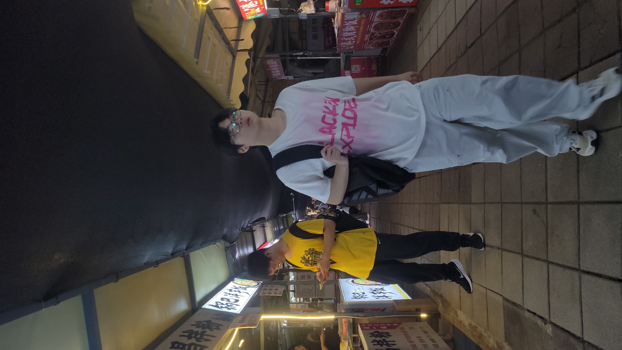 The image, rotated 90 degrees clockwise, depicts a nighttime scene on a busy urban street or market. Two young individuals stand on a paved sidewalk, illuminated by artificial lights from surrounding stalls.
The person in the foreground wears glasses, a white short-sleeved t-shirt with pink text "LACK XPLODE" (partially visible), light-colored loose pants, and white and black athletic shoes. A black shoulder bag is slung across their body, and they are looking slightly upwards and to their right.
Behind them, another person is visible, wearing a yellow short-sleeved t-shirt with a dark design, dark pants, and black athletic shoes with white soles. This individual appears to be holding a phone or small object, looking down at it.
To the right, a row of stalls or small shops line the street, featuring bright, illuminated signs with Chinese characters and images of food. Visible text on various signs includes "元", "拌粉销", "10元", "饼价", "商品", and "重庆快". The ground is paved with square tiles. The scene conveys the atmosphere of a lively evening street food or shopping area.