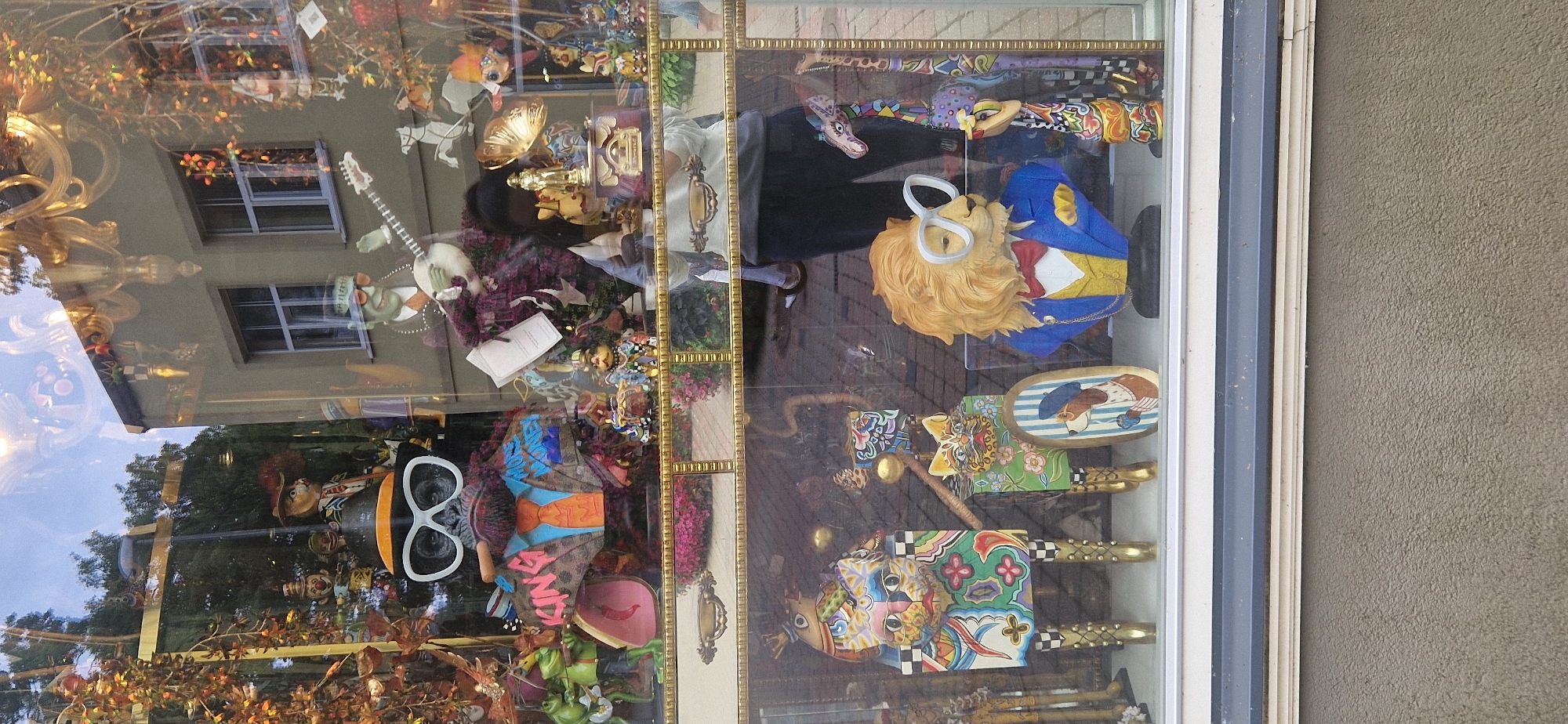 The image shows a colorful and eclectic display in a shop window. In the foreground, there are several whimsical figurines. To the left, a large, stylized cat statue with a multi-colored, patterned body and gold legs stands prominently. Next to it, a smaller cat statue with similar artistic flair is positioned. In the center, a bust of a lion wearing large white glasses, a blue jacket, a yellow vest, and a red bowtie is displayed. To the right, a tall, colorful giraffe figurine stands. Further back, a bust of a gorilla wearing a hat, glasses, and a suit with "KING" and "MORE MONEY" written on it is visible. There is also a figure of a green frog, a man playing a banjo, and various other decorative items. The window has reflections of trees, buildings, and the sky, suggesting it is an outdoor display. The overall impression is one of a quirky and artistic collection of decorative objects.
