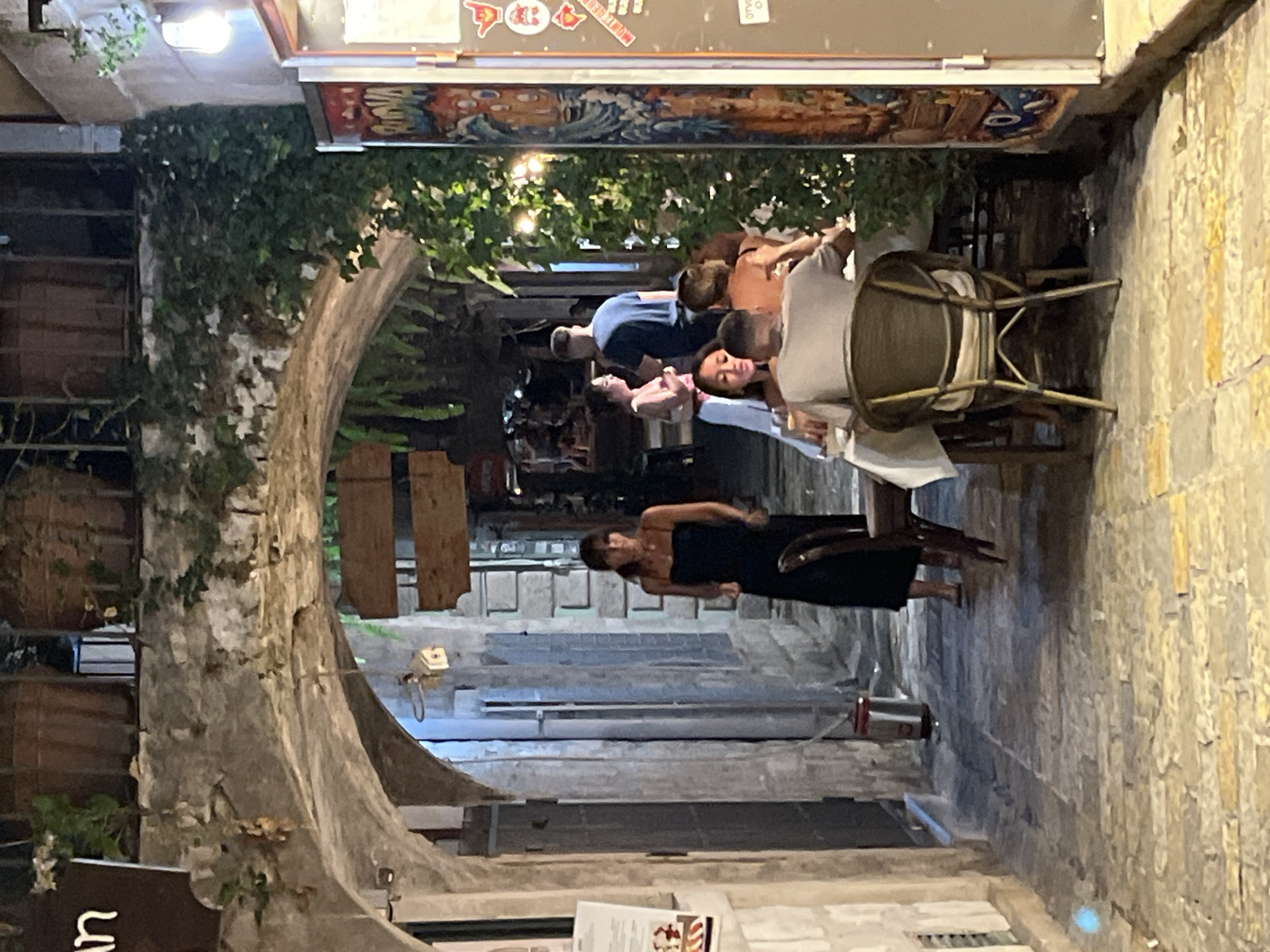 In Budva, Montenegro, an outdoor dining area is situated under an arched stone passageway, illuminated by ambient lighting. The scene features several individuals engaged in dining and conversation. A woman in a dark dress stands to the left, while a group is seated at a table covered with a white tablecloth in the mid-ground. Two men are visible at this table, one facing away from the camera, and another partially obscured. Further back, a couple stands near a brick wall, the woman in a light-colored dress and the man in a blue t-shirt. The passageway is paved with stone and bordered by old stone buildings and overgrown greenery. A colorful mural or advertisement adorns a structure on the right side of the frame, and a trash receptacle is visible to the left.