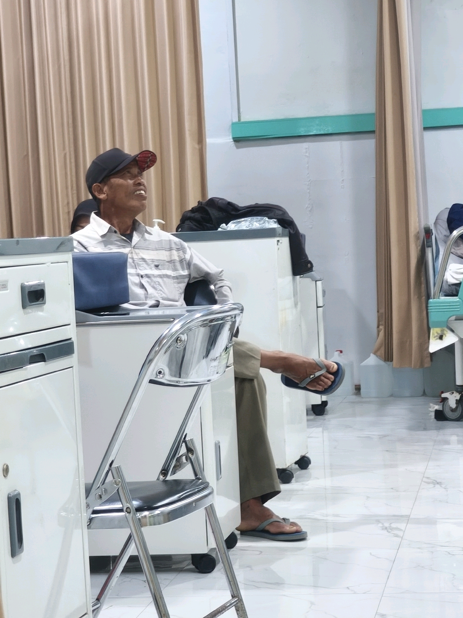 A man wearing a black baseball cap and a light-colored, striped button-down shirt sits in a silver folding chair next to a white medical cart. He is looking upwards with his mouth open, as if speaking or in distress. His right leg is extended, and he is wearing khaki pants and blue flip-flops. Another person is partially visible behind him. The scene appears to be indoors, possibly a hospital or clinic waiting area, with tan curtains in the background and a white tiled floor. The overall impression is of a moment captured in a public space.