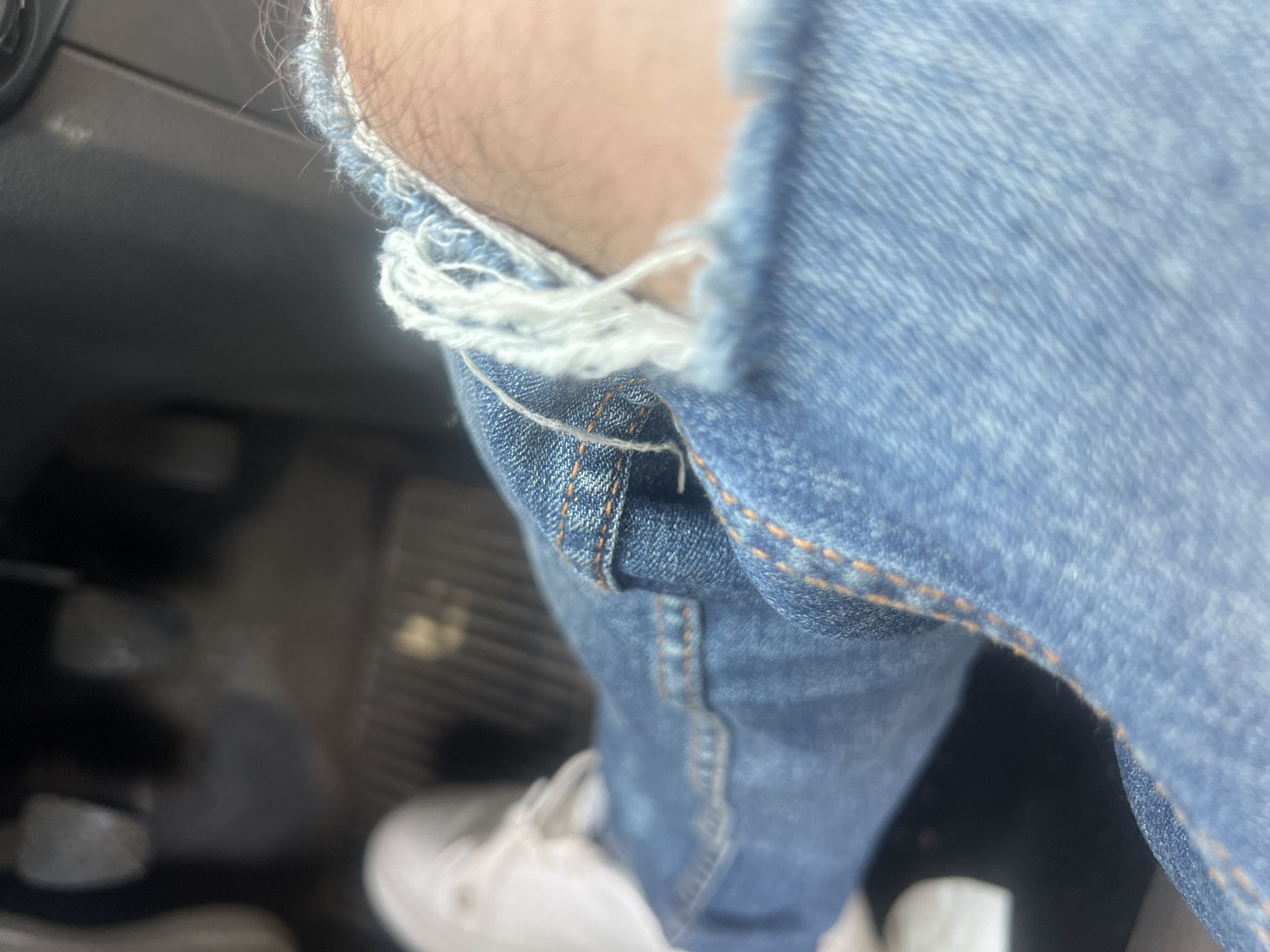 The image displays the lower half of an individual seated within a vehicle in Casablanca, Morocco. The subject is wearing light blue ripped denim jeans, with a prominent tear on the left leg revealing exposed skin and frayed white threads. White athletic shoes are visible on the feet, positioned near the vehicle's floor. In the blurred background, three distinct pedals typical of a manual transmission vehicle are present: a clutch pedal on the far left, a central brake pedal, and an accelerator pedal on the right. A dark, ribbed floor mat or carpet is visible beneath the pedals. The dark gray interior trim of the car's dashboard or console is visible in the upper right portion of the frame. The scene is illuminated by ambient light, suggesting a daytime setting.