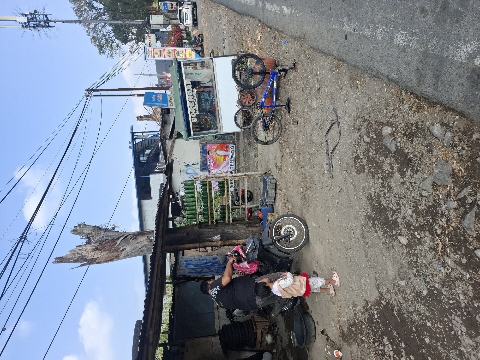 The image depicts an outdoor scene on a roadside in Sukabumi, Indonesia. In the foreground, a man in a black t-shirt is attending to a black motorcycle, which is parked on a dirt and gravel surface. A young girl in a light-colored jacket and red skirt stands beside him, looking at the motorcycle. To the left of the motorcycle, there is a large black tub and a stack of tires. Behind the man and motorcycle is a simple open-sided shelter with a corrugated metal roof, supported by wooden posts. A wooden shelf unit filled with green glass bottles, likely fuel, is visible behind the motorcycle.
In the middle ground, a food cart with a green awning and the word "GORENGAN" (fried food) written on it is present. A bicycle, blue and black, is disassembled and lying on the ground near the food cart, with parts including wheels and a frame visible. Signs and posters are affixed to structures in the background. One poster features a man's face and text in Indonesian. Further back, a white building with a blue roof is visible, and beyond that, a cell tower with multiple antennas stands against a bright blue sky with scattered white clouds. The road runs along the right side of the image, with a white car parked further down. The overall environment suggests a somewhat unkempt roadside area with commercial activity, typical of many urban or semi-urban settings in Indonesia.