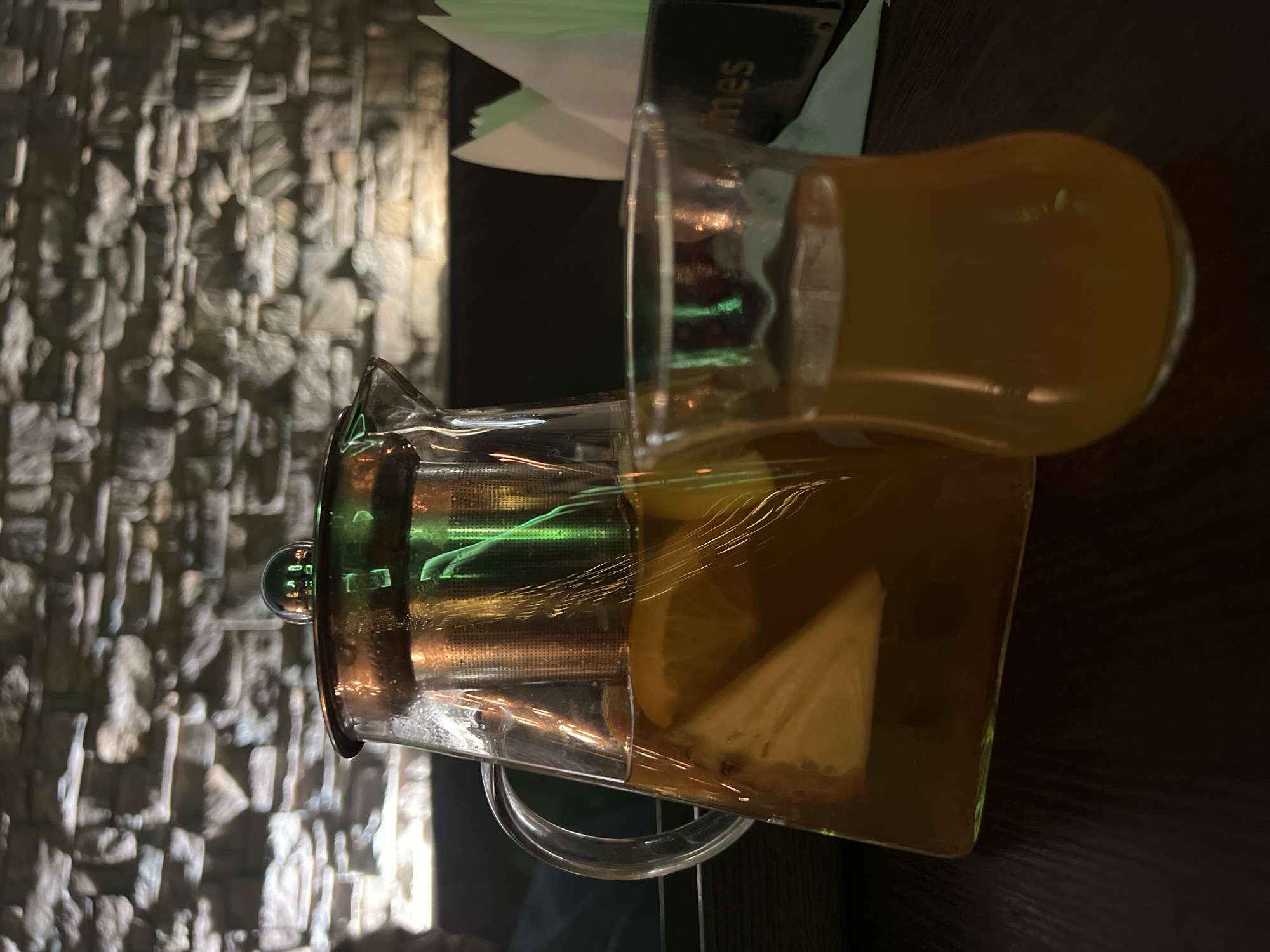 A glass pitcher with a built-in strainer and a glass cup are on a table, likely indoors. The pitcher contains a yellow liquid, possibly tea, with lemon slices visible. The cup is filled with the same liquid. The table is dark, and the background shows a textured stone wall. The location in the city of Zaosinovo, Russia is not discernible from the image.