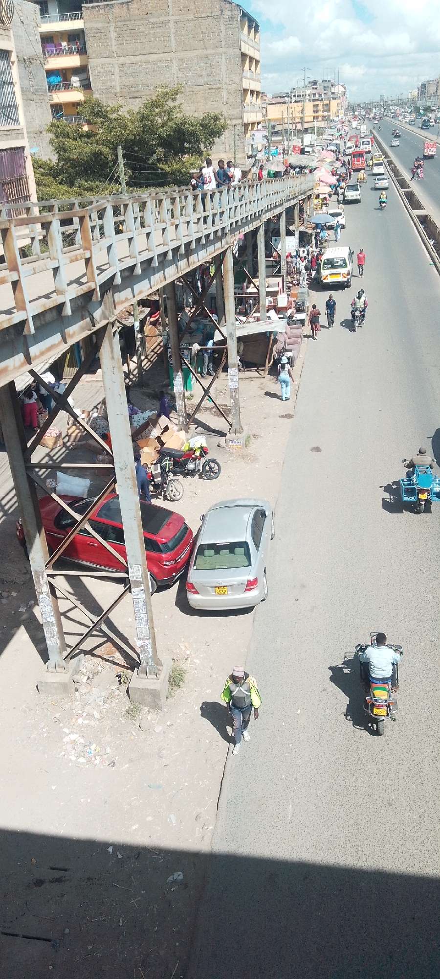 The image shows an elevated view of a bustling street scene in Nairobi, Kenya. A pedestrian footbridge spans a major thoroughfare filled with cars, vans, motorcycles, and three-wheeled vehicles. People are on the footbridge and clustered beneath it, where makeshift stalls and merchandise are displayed. Two cars, one red and one silver, are parked underneath the bridge structure. A man wearing a hat and a green vest walks along the roadside next to a motorcyclist. The surrounding area includes multi-story buildings, some with visible balconies. Traffic is heavy on the road.