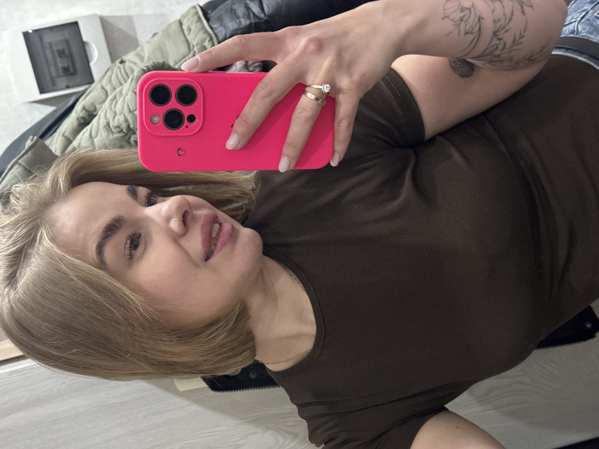 A person is holding a pink smartphone to take a selfie. The person has blonde hair, and the skin of their right arm shows a floral tattoo. They are wearing a brown t-shirt and a ring on the third finger of their left hand. Background details include what appears to be a jacket and a white box. There is no indication of the location being Chaikovsky, Russia in the provided image.