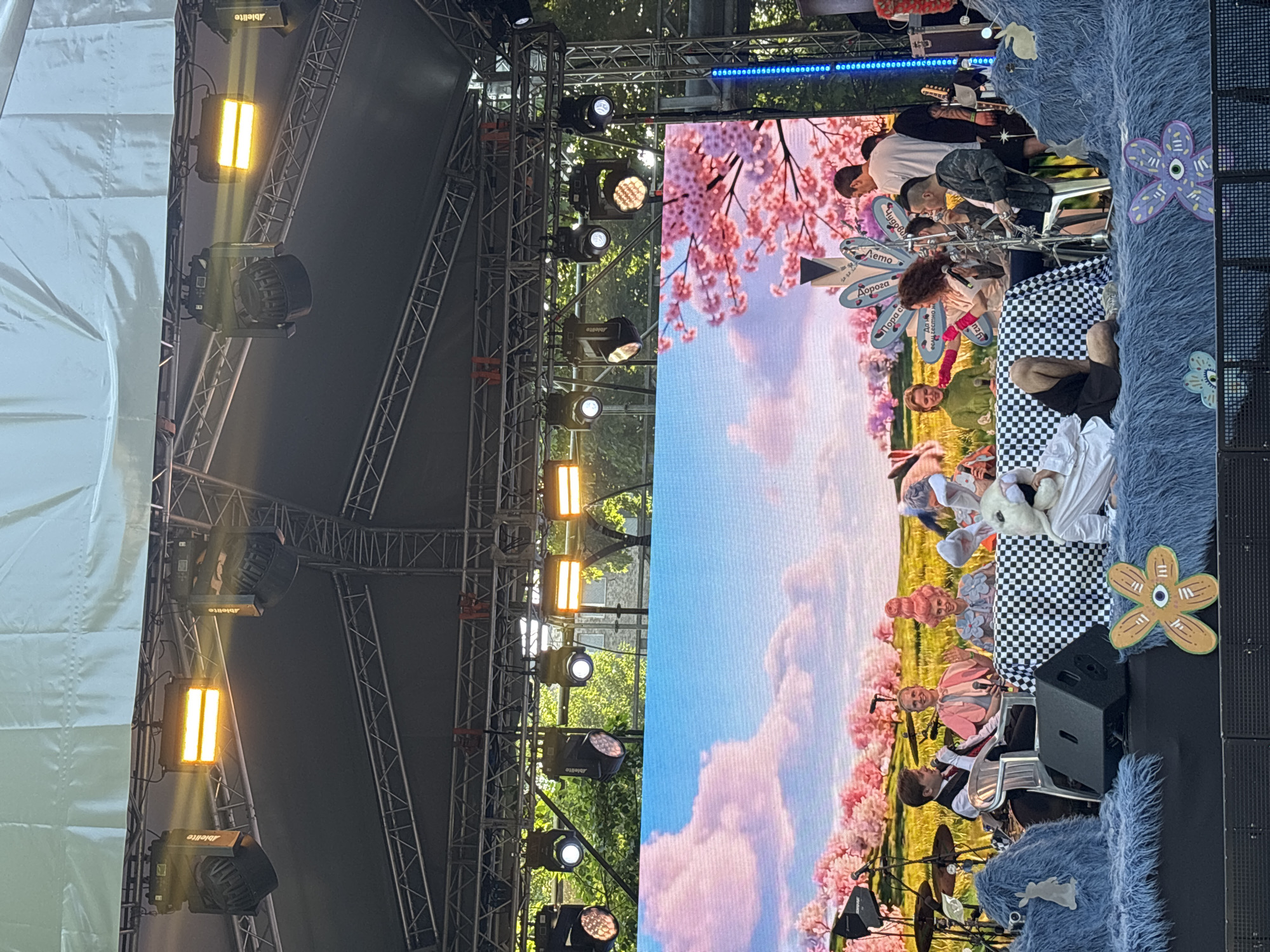 The image shows a stage setup in an outdoor setting, likely in Moscow, Russia. The stage is equipped with lighting rigs and a large screen displaying a pastoral scene with cherry blossoms and a cloudy sky. On the right side of the stage is a seating area with a checkered pattern. Multiple performers are on the stage, some playing instruments, and others seated. Decorations such as flowers, and stuffed animals can be seen on the stage. The environment indicates a performance or event taking place outdoors with a large stage for the entertainers.