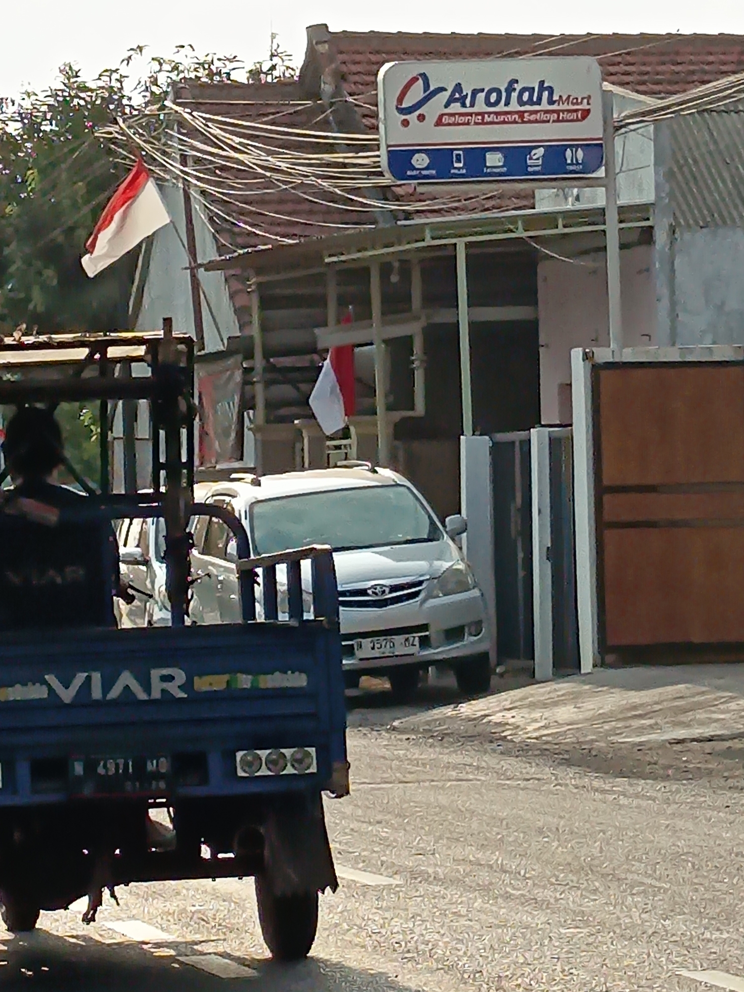 A low-angle, medium shot captures a blue three-wheeled truck parked on a paved street in front of a commercial building. The truck's rear is visible, showing the brand name "VIAR" and license plate "N 4971 MO". Three circular lights are mounted on the back of the truck. To the right of the truck, a silver car is parked, partially obscuring the entrance to a building. The building features a red-tiled roof and a prominent sign for "Arofah Mart" with smaller icons below. Indonesian flags, red and white, are displayed on poles near the building. The environment suggests a sunny day in a semi-urban or rural area, possibly a small business district. The background shows trees and a glimpse of what appears to be a residential structure.