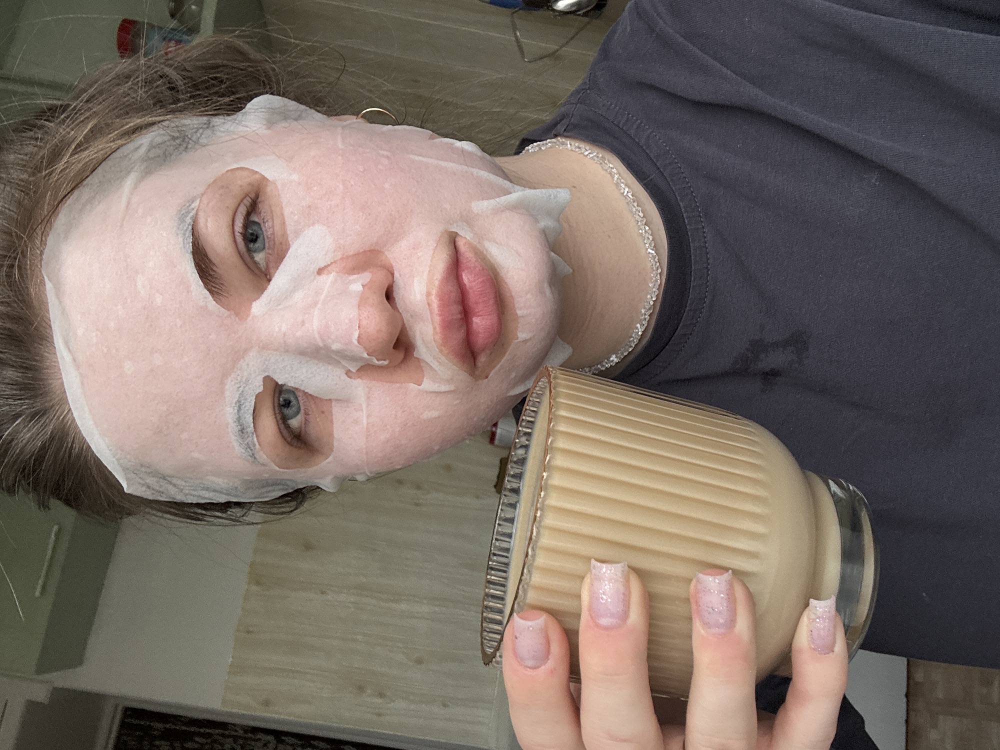 A person wearing a white facial mask is holding a glass of light brown liquid. The person has blue eyes and is wearing a dark gray shirt with a silver-colored necklace. The nails on the hand holding the glass are painted with a light pink glitter polish. The background appears to be a domestic indoor environment with light-colored walls and a partially visible floor. There is no verifiable evidence in the image to confirm the location is Chaikovsky, Russia.