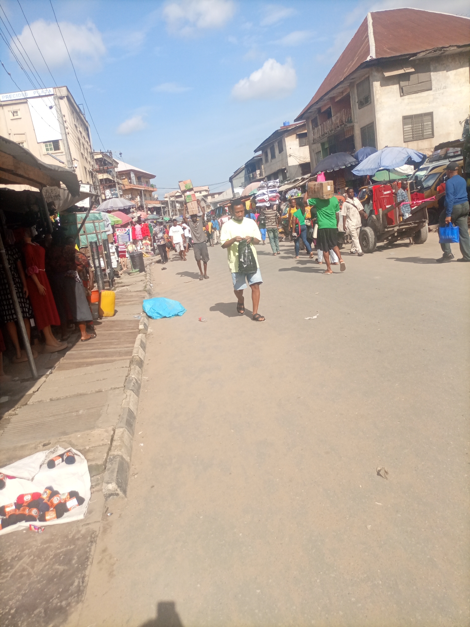 This is an outdoor, eye-level, medium shot of a bustling street scene in Fegge, Nigeria. The sun is shining, casting shadows on the pavement, and the sky is a pale blue with scattered white clouds. The street is paved and appears to be a market area with various stalls and shops lining one side.
In the foreground, a man in a yellow shirt and blue shorts walks towards the viewer, carrying a black plastic bag. To his left, a blue plastic bag is discarded on the ground, next to a small bottle. Further left, along the side of the street, a market stall is set up with mannequins displaying clothing, including red and black dresses. A yellow plastic container sits on the wooden walkway in front of the stall.
In the middle ground, the street is filled with people walking and interacting. Several individuals are carrying boxes or bags of goods on their heads or shoulders, suggesting a busy marketplace. A red and white cart with large wheels is visible, with people gathered around it. Umbrellas are set up over some of the stalls, providing shade.
The background features buildings that appear to be multi-story structures, some with balconies and corrugated metal roofs. The architecture suggests an urban environment. The overall impression is one of a lively, everyday commercial scene in a Nigerian city.