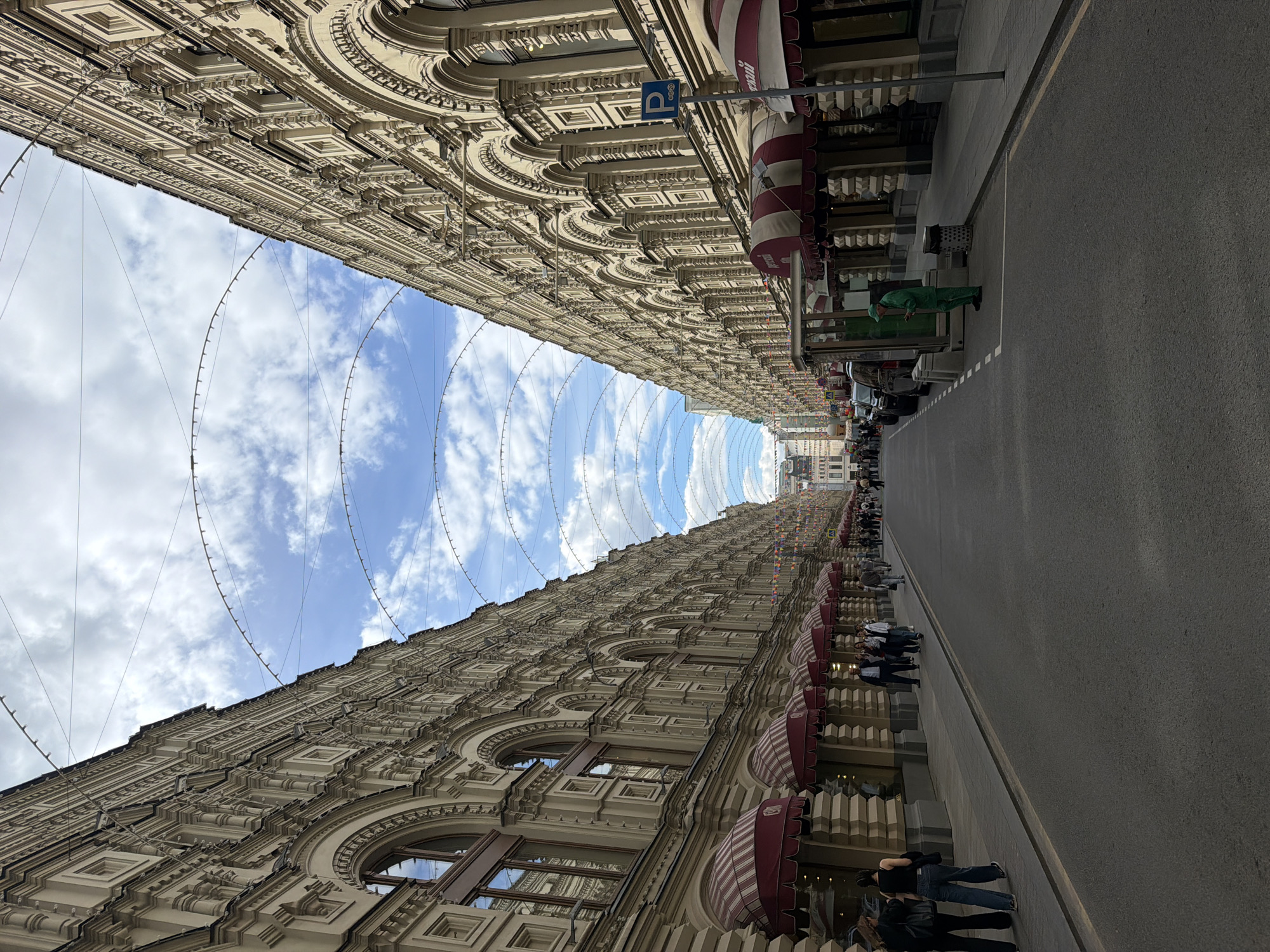 The image depicts a street scene in what appears to be a city with classical architecture. The buildings lining the street are tall, ornate, and feature intricate detailing. A series of suspended, semicircular light installations span across the street, creating a unique visual effect against a backdrop of blue sky and scattered clouds. On the right side of the image, pedestrians are visible walking along the sidewalk and street. Storefronts with striped awnings line the base of the buildings. A parking sign is attached to the building. A man in green scrubs can be seen on the right. The street is paved and appears to be well-maintained. The image is oriented vertically.
