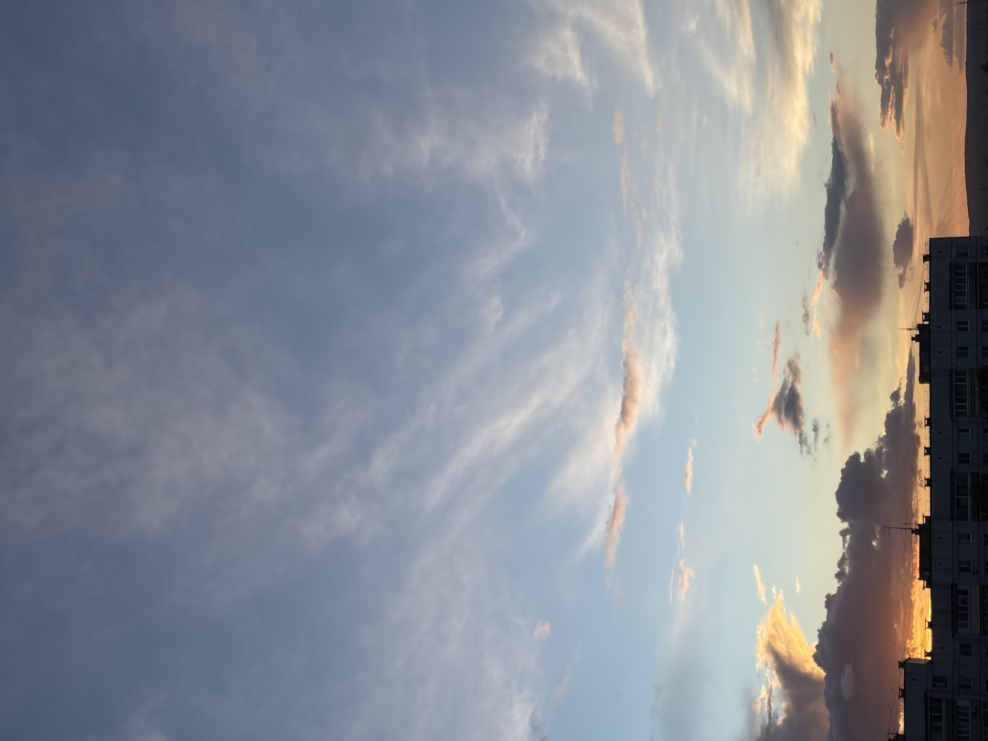 The image captures a view of the sky at dusk or dawn in Chaikovsky, Russia, with a predominantly blue sky on the left transitioning to lighter blues and oranges on the right. Wispy clouds are scattered throughout the frame. Towards the right, the horizon features a building, possibly an apartment block, its silhouette defined against the colorful sky. The clouds near the horizon display a mix of grey and orange hues, indicative of the sun's position just below the horizon line. Power lines can be seen running along the top of the building.
