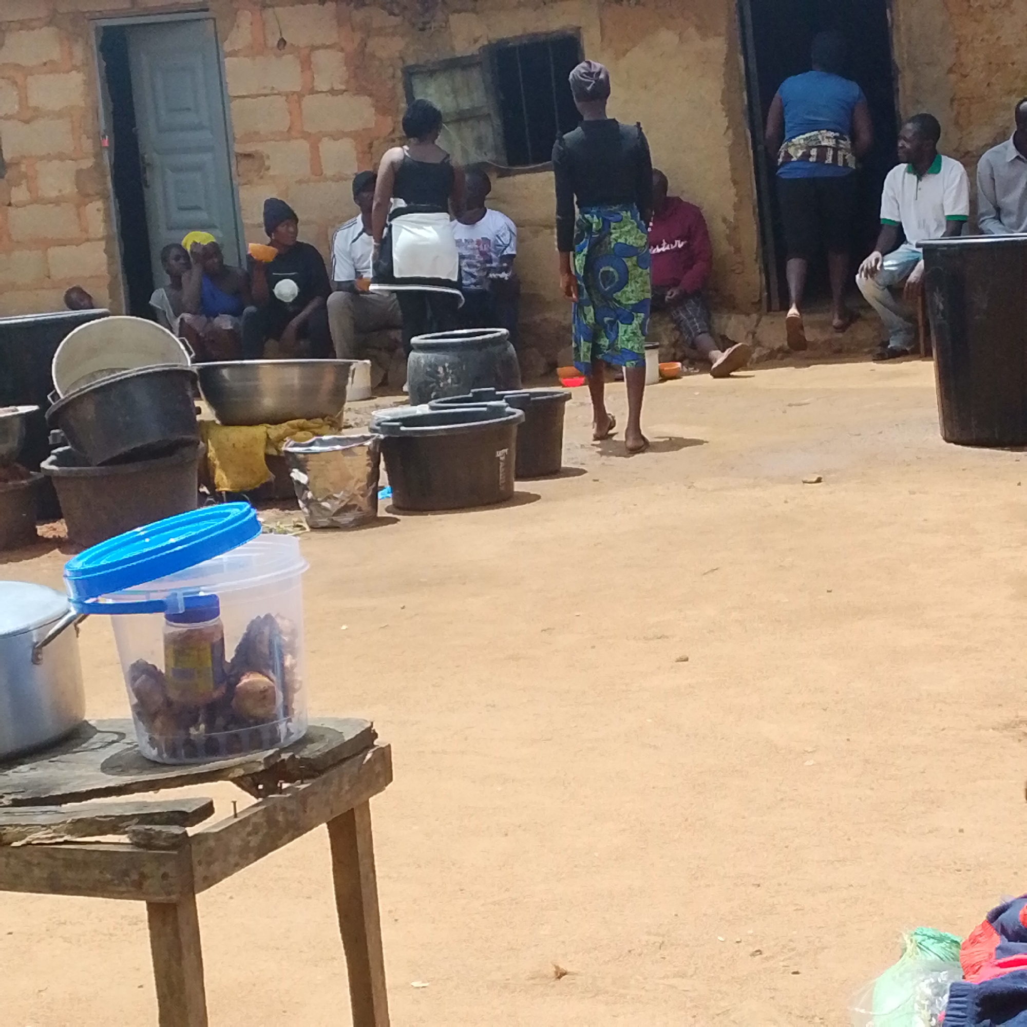 The image depicts an outdoor scene in what appears to be a village or rural area. The backdrop consists of a building constructed from rough, light-colored bricks. A doorway with a gray, paneled door is visible on the left side of the building. The ground is covered in reddish-brown dirt.
Several individuals are present in the scene. On the left, a group of people are seated near the doorway. Further to the right, two women are standing, one wearing a white apron over a black dress and the other a black top and a colorful patterned skirt. Several men are also seated or standing in various parts of the image. One man in the center is wearing a white polo shirt with green trim and light-colored pants. Another man on the far right is wearing a light gray shirt. A man on the far left is wearing a red hoodie.
A variety of objects are scattered around the scene, predominantly containers. These include large black plastic tubs, metal pots, and a clear plastic container holding some dark objects and a small jar. A broken wooden table is positioned in the foreground on the left. The overall impression is one of a communal gathering or a household engaged in activities, possibly related to cooking or food preparation. The lighting suggests it is daytime, with shadows indicating the sun's position.