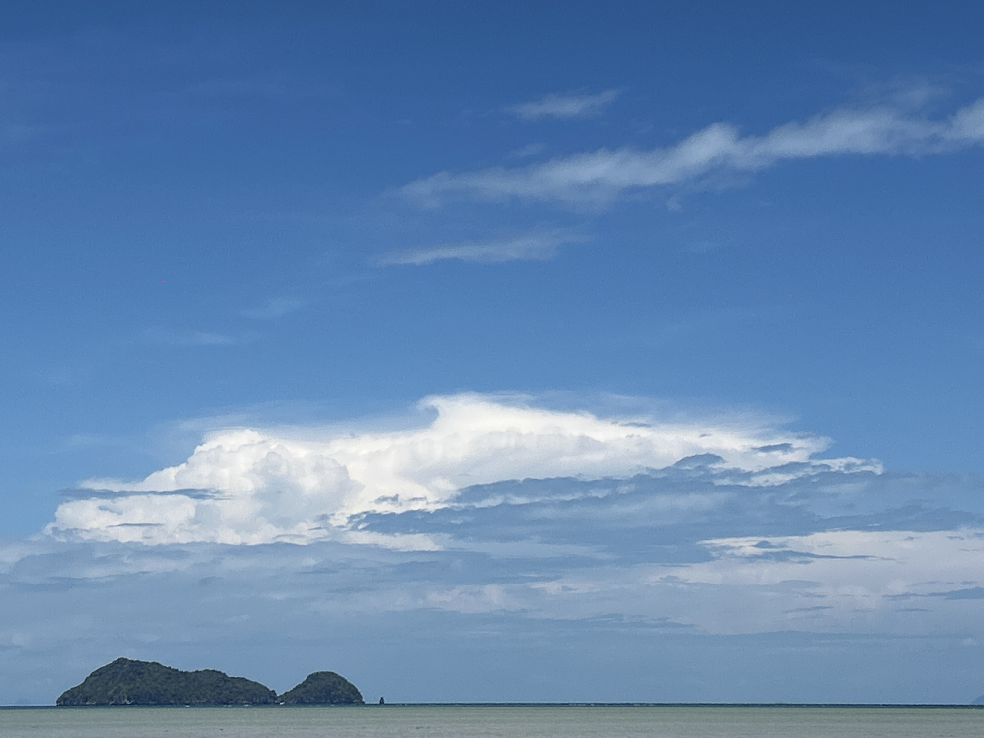 The image shows a seascape in ตำบลเกาะพะงัน, Thailand. The sky is predominantly blue with scattered clouds, including a large, puffy cumulus cloud formation in the middle. Below the sky, a calm, light-colored sea stretches to the horizon. Two small, green islands are situated near the center of the frame, in the distance.