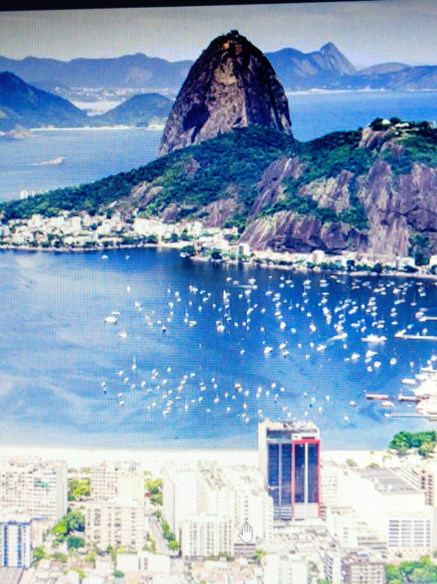 This is a high-angle, full shot of a city skyline adjacent to a body of water with mountains in the background. The city has a dense collection of buildings, including high-rise apartments and what appears to be an office building with red accents in the foreground. The water is filled with numerous boats, mostly sailboats. The background features lush green mountains, one of which is a prominent, rocky peak. The sky is a bright, hazy blue with a few white clouds scattered throughout. The image appears to be a photograph, possibly taken from a drone or a very tall building.