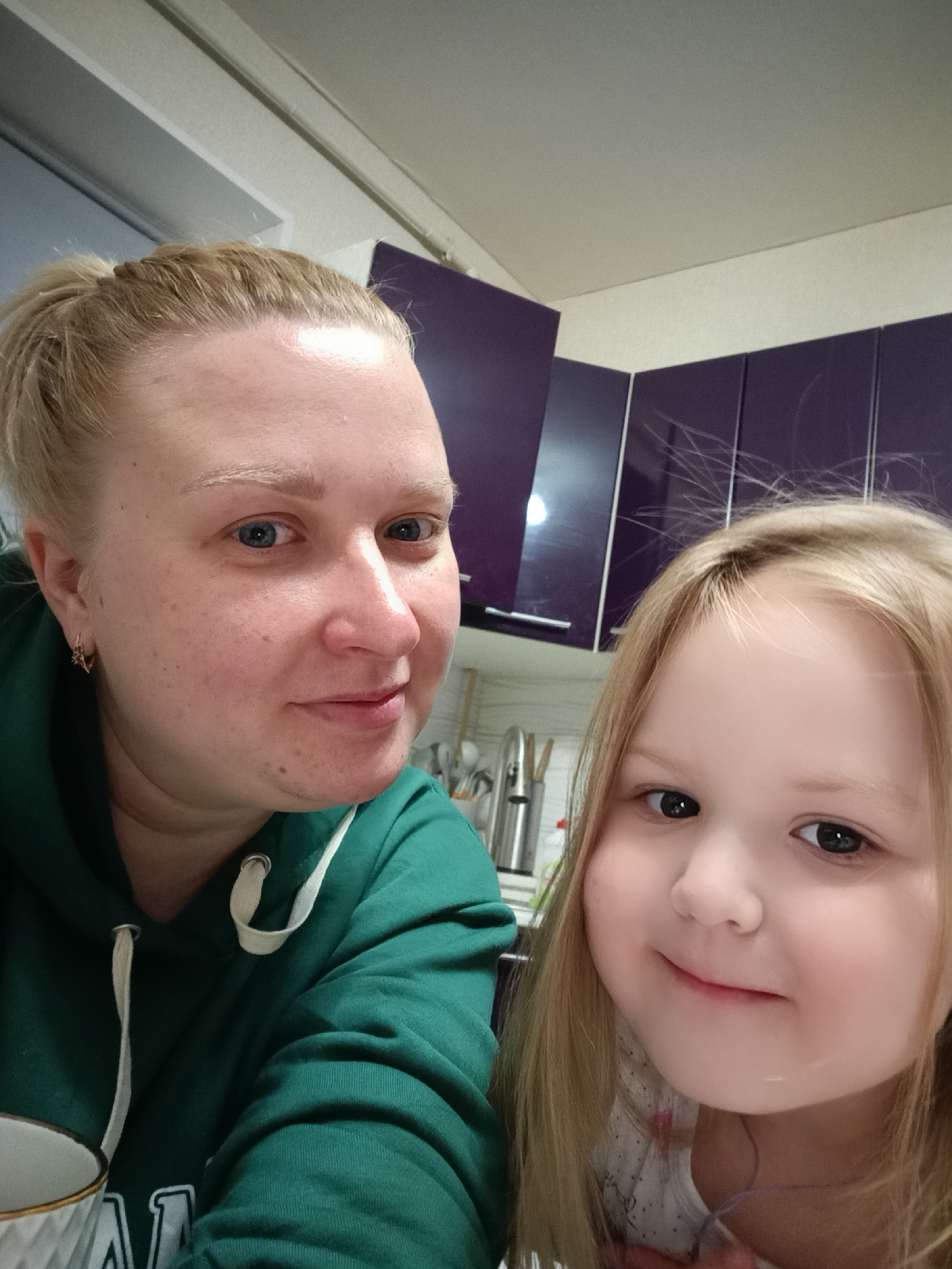 The image shows a selfie of a woman and a young girl. The woman is on the left, wearing a dark green hoodie with white drawstrings. Her blonde hair is pulled back. The girl is on the right with long, light brown hair. The background shows what appears to be a kitchen setting, with dark purple cabinets and a white countertop visible. A stainless steel faucet and utensils can also be seen. The location is likely in Chaikovsky, Russia, based on prior knowledge.