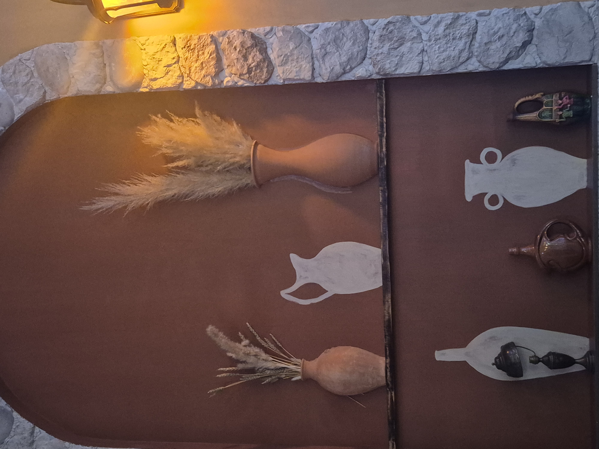 The image depicts a rustic display shelf with various decorative items set against a reddish-brown wall, framed by a stone archway. The shelf holds several vases and decorative objects. On the left, a terra cotta vase is filled with dried grasses and wheat stalks. Next to it, a white silhouette of a pitcher is painted on the wall. Further to the right, a tall, slender terra cotta vase contains a large bouquet of fluffy, dried reeds.
Below this, on a lower shelf, a tall, flat, white vase-like object is displayed, featuring a miniature antique-style lamp with a dark metal base and shade. To its right, a brown ceramic jug with a curved handle and decorative etching is visible. To the far right on the lower shelf, a small, multi-colored decorative item with what appears to be a painted scene is present, along with a white, amphora-shaped vase with two loop handles. A warm yellow light emanates from a lamp to the right of the scene, casting a glow on the stone archway and the wall. The overall ambiance suggests an interior decorative setting. There is no information available to determine the exact location within Kokshetau, Kazakhstan.