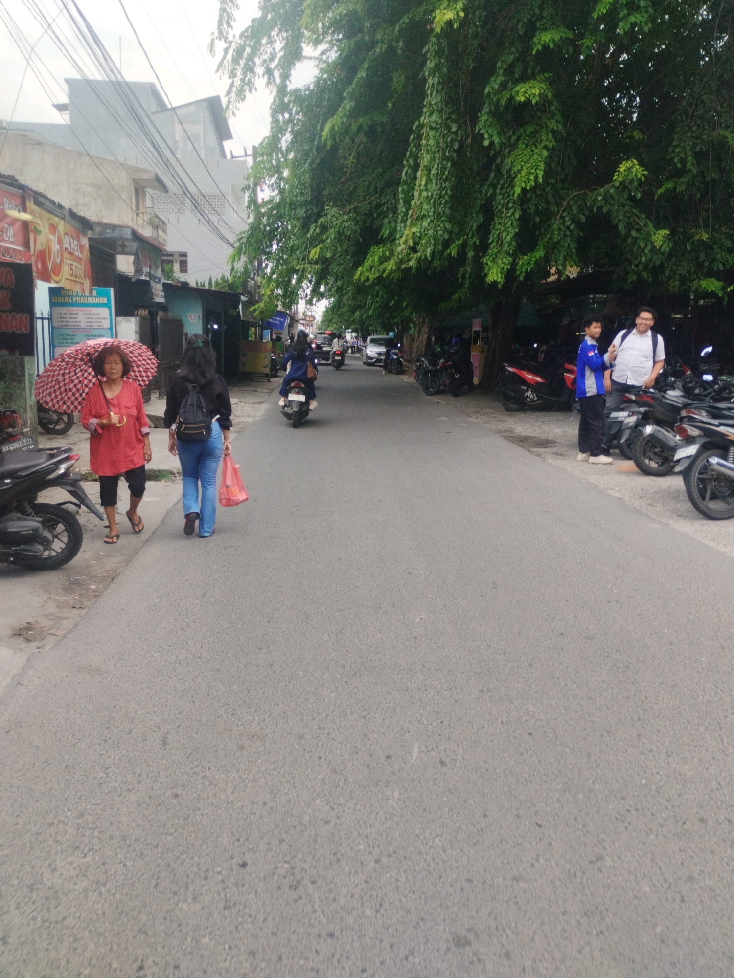 An outdoor, eye-level, medium shot captures a street scene in the City of Medan, Indonesia. The image depicts a moderately wide asphalt road with a sidewalk on the left. A large tree with dense green foliage overhangs the right side of the road, casting shadows. Several motorcycles are parked along the right side of the road, and a few people are visible.
On the left sidewalk, a woman with short, dark hair, wearing a red long-sleeved shirt and dark pants, walks towards the viewer while holding a red and white checkered umbrella. Next to her, walking away from the viewer, is another woman wearing a black long-sleeved shirt, blue jeans, and carrying a black backpack. She holds a red plastic bag in her right hand. A black motorcycle is parked on the far left of the sidewalk.
Further down the road, a person on a motorcycle rides away from the viewer. On the right side of the road, two individuals stand near the parked motorcycles. One is a young person wearing a blue and white jacket and dark pants, and the other is an adult male wearing a white short-sleeved shirt and dark pants, with a backpack slung over his left shoulder.
Buildings are visible in the background on the left, including a multi-story structure with a light-colored facade and exposed electrical wires running across it. A sign with Indonesian text and numbers is attached to one of the buildings. The overall environment appears to be a typical urban street with shops and residential buildings lining the road. The sky is overcast.