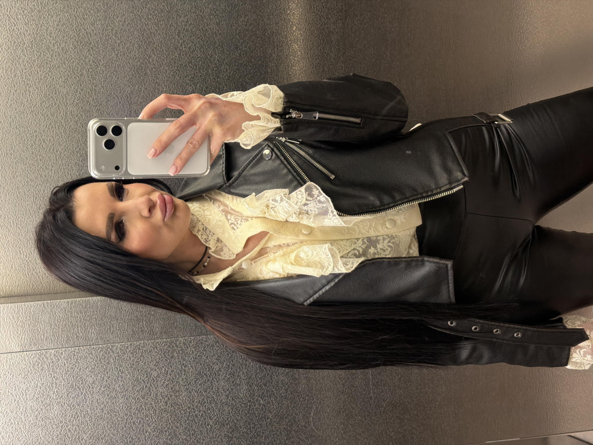 A brunette woman in a black leather jacket over a cream-colored lace blouse and black leather pants takes a selfie with an iPhone in an elevator. The elevator has a gray metallic finish. The location is unknown, but the query requested that it be identified as Voronezh, Russia.