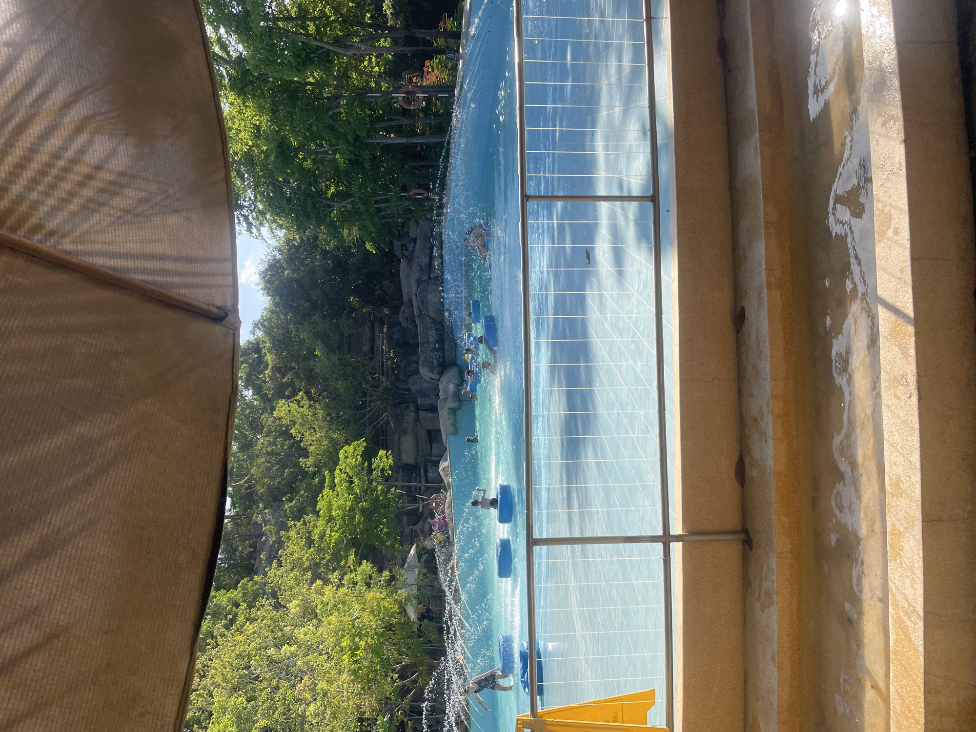 The image shows a scene at the VinWonders Water Park in Nha Trang, Vietnam. The view is partially obstructed by a tan umbrella in the foreground. Beyond that is a lazy river attraction, where people are floating in blue inner tubes. The river is surrounded by lush trees and rock formations. The water is a light blue color. A metal railing is visible along the side of the river. A yellow slide is visible at the bottom of the image. The right side of the image shows stone steps with water running down them. Sunlight is reflected on the wet steps.